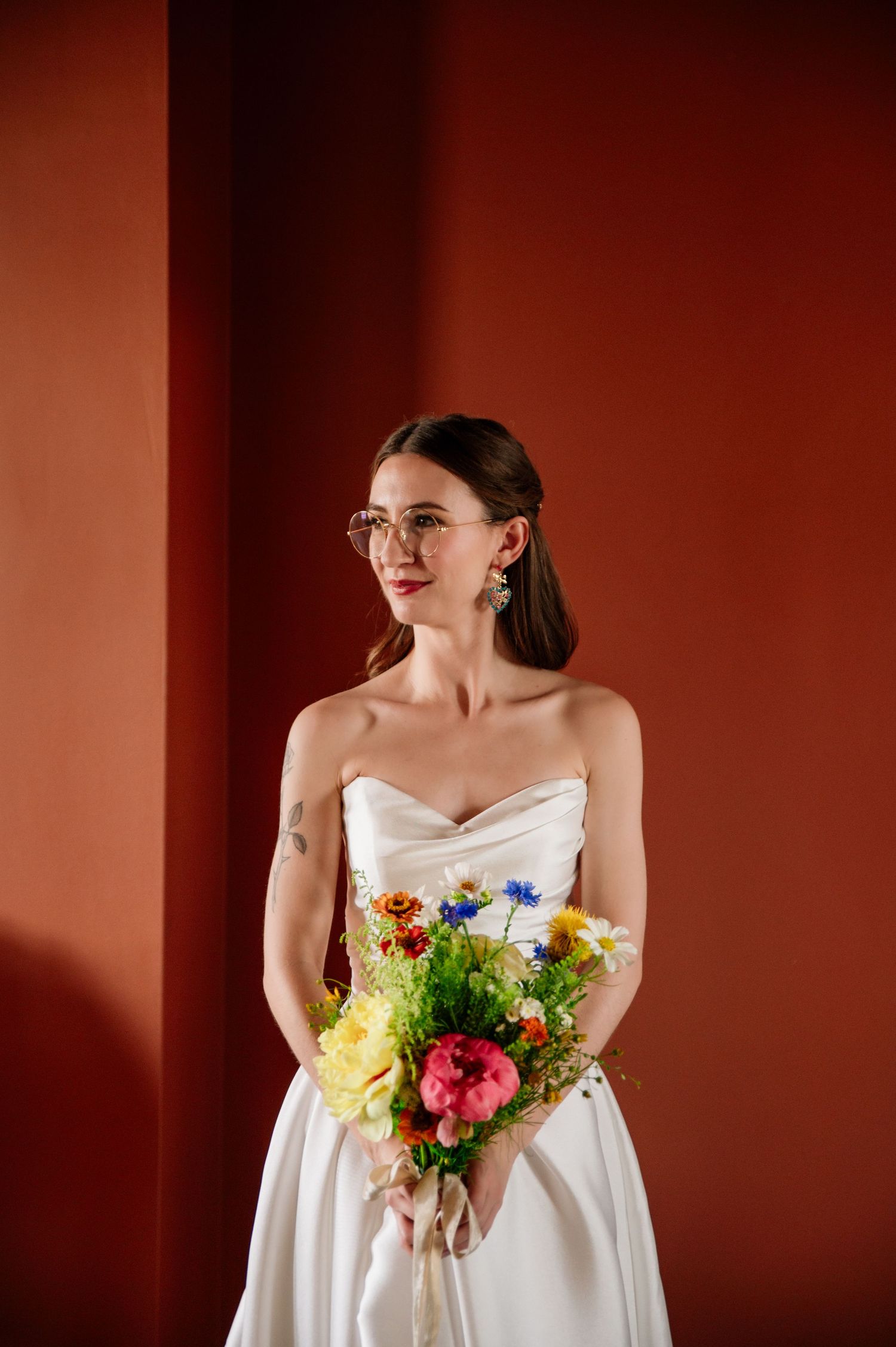 A portrait of someone wearing a white strapless wedding dress holding a colorful bouquet against a red background.