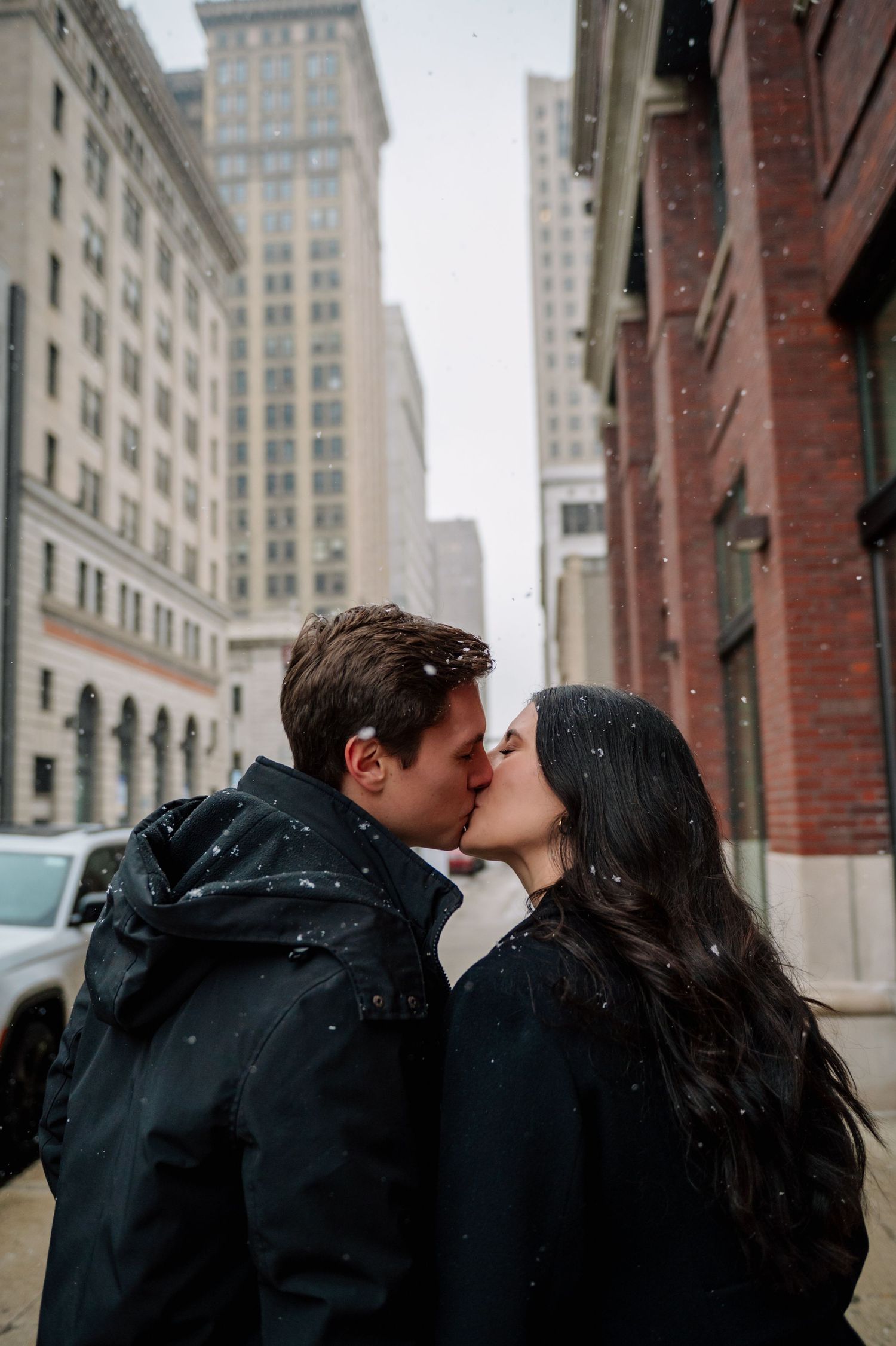 A couple shares a romantic kiss on a snowy day in the city with tall buildings in the background.