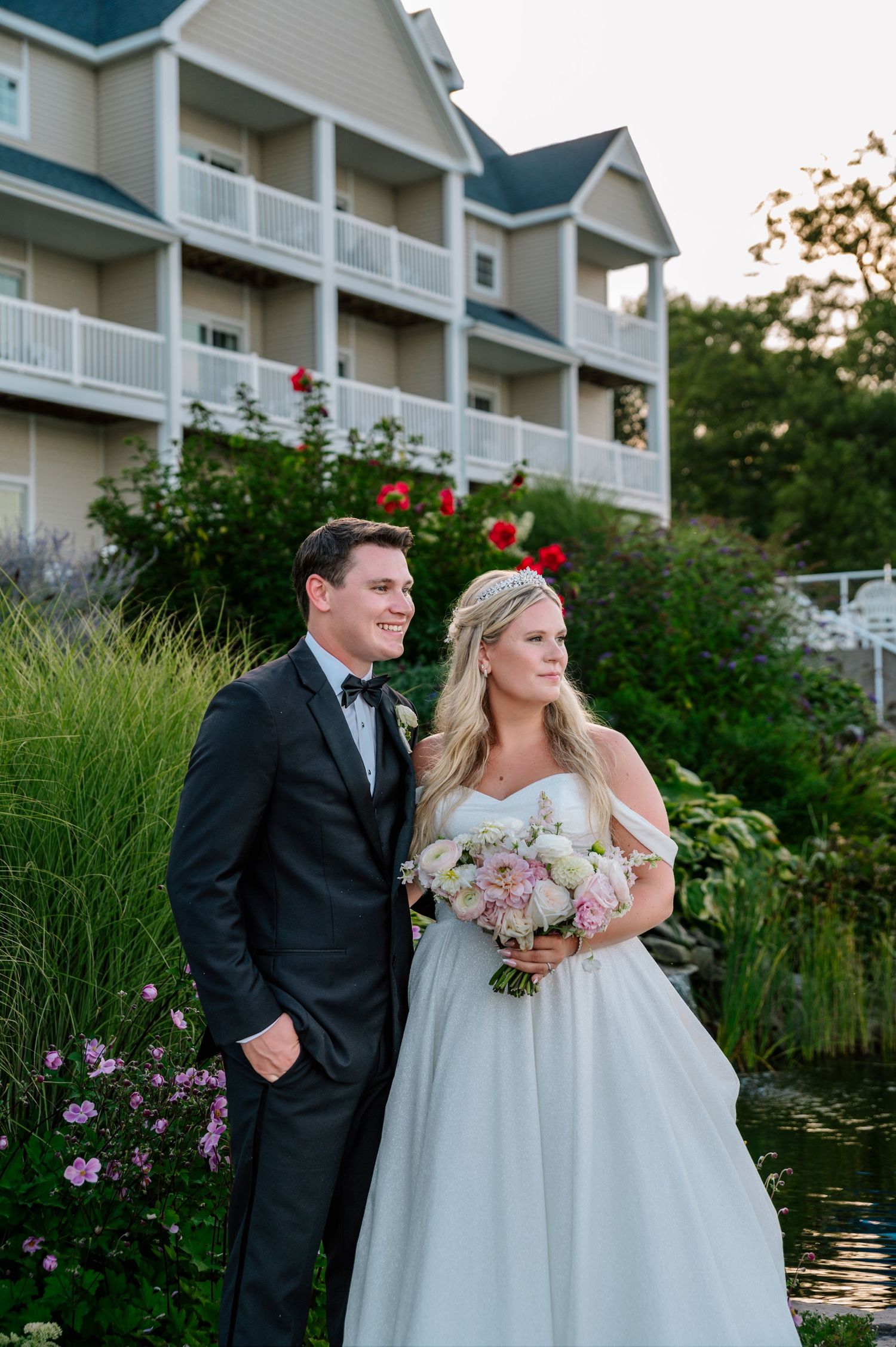 Elegant wedding couple poses by a waterfront garden with white building and flowers in the background.