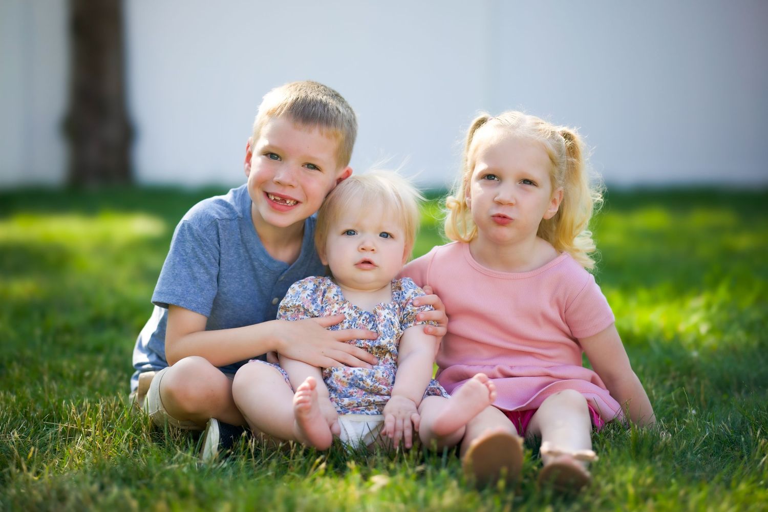 Three young children sit together on green grass during a sunny summer day outdoors.