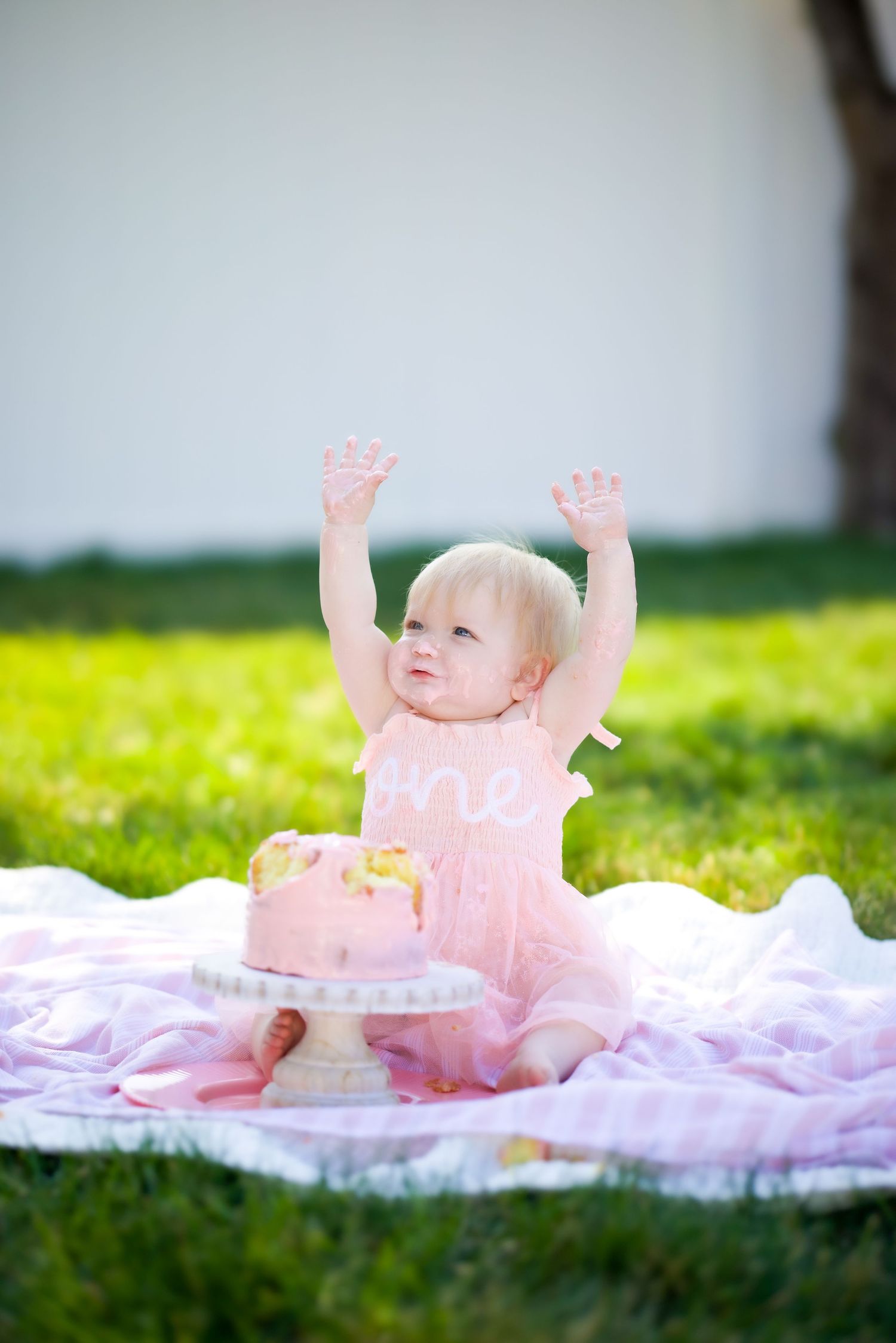 A baby in a pink dress raises their arms joyfully while sitting on a blanket with a birthday cake outdoors on green grass.
