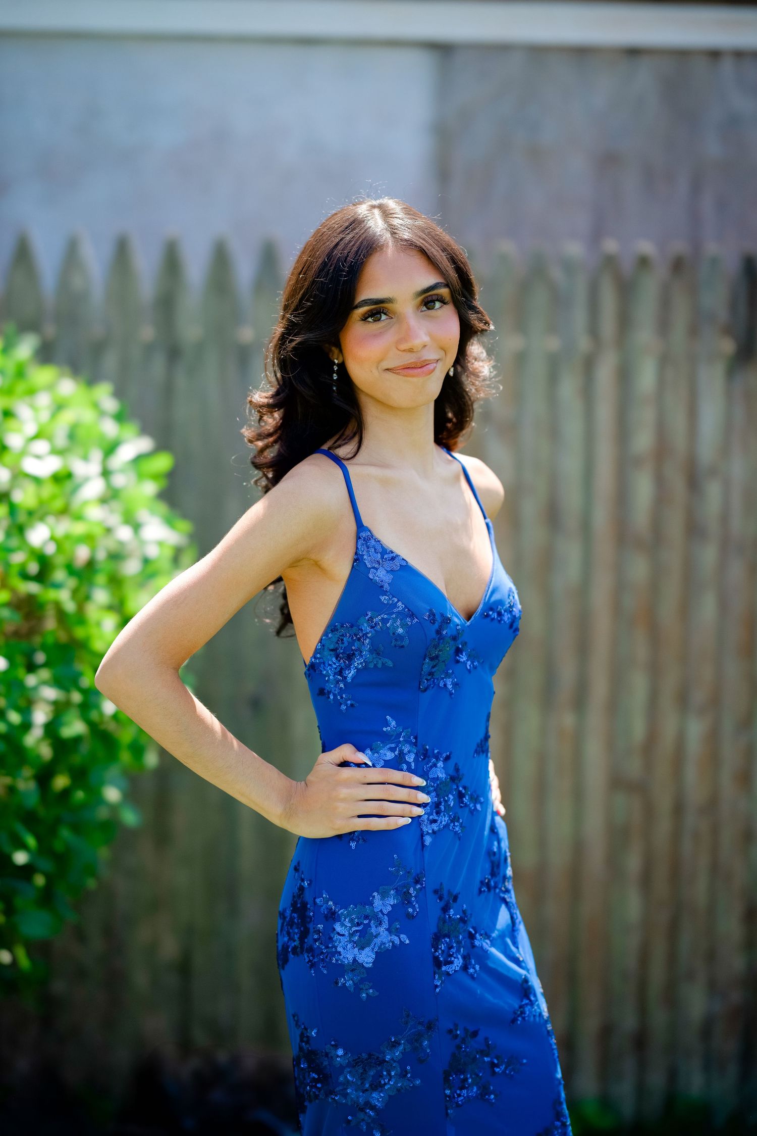 A woman in an elegant royal blue lace dress poses outside in a garden setting against a wooden fence.