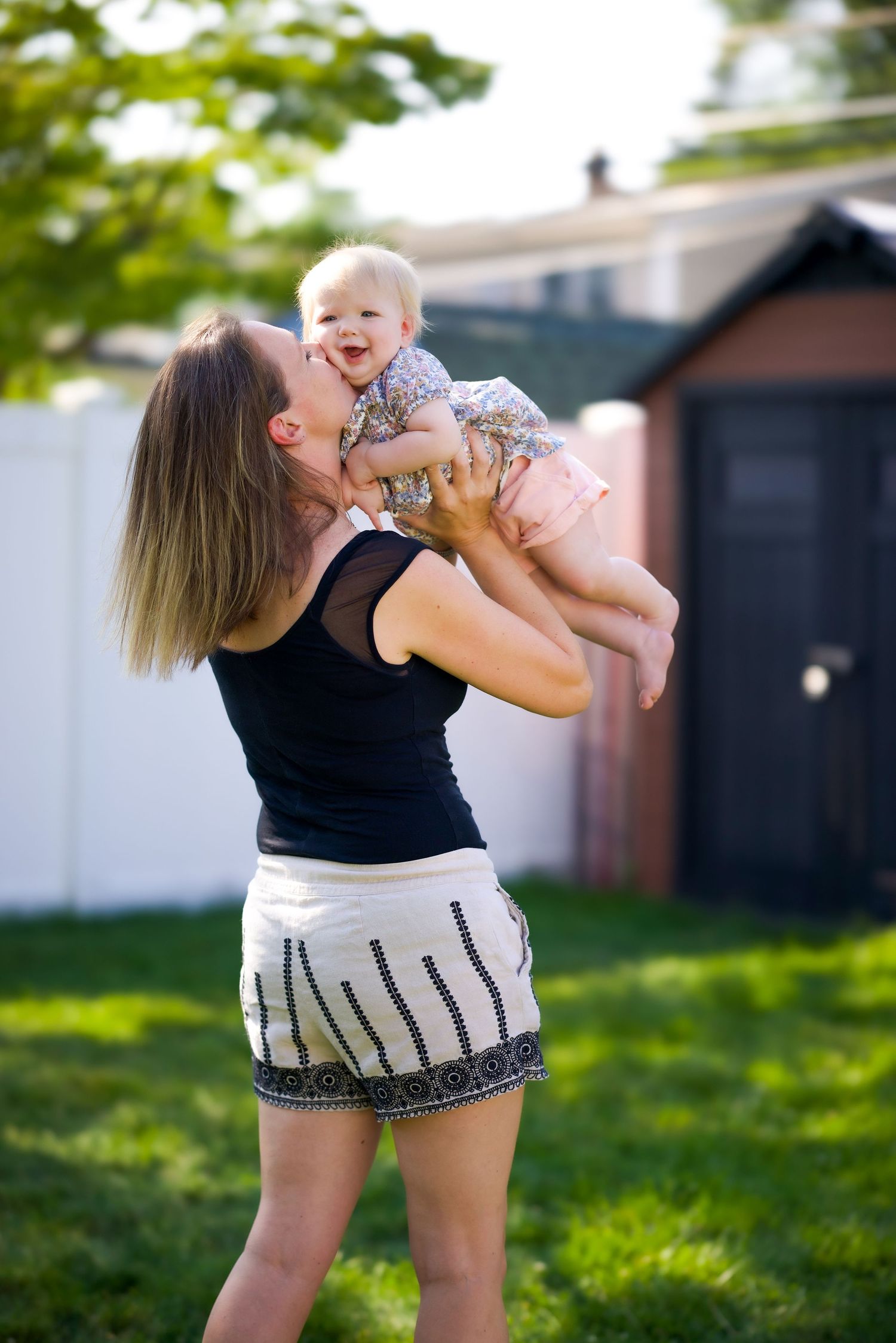 A parent in a black tank top and patterned shorts lifts a laughing baby in a sunny backyard with a shed in background.