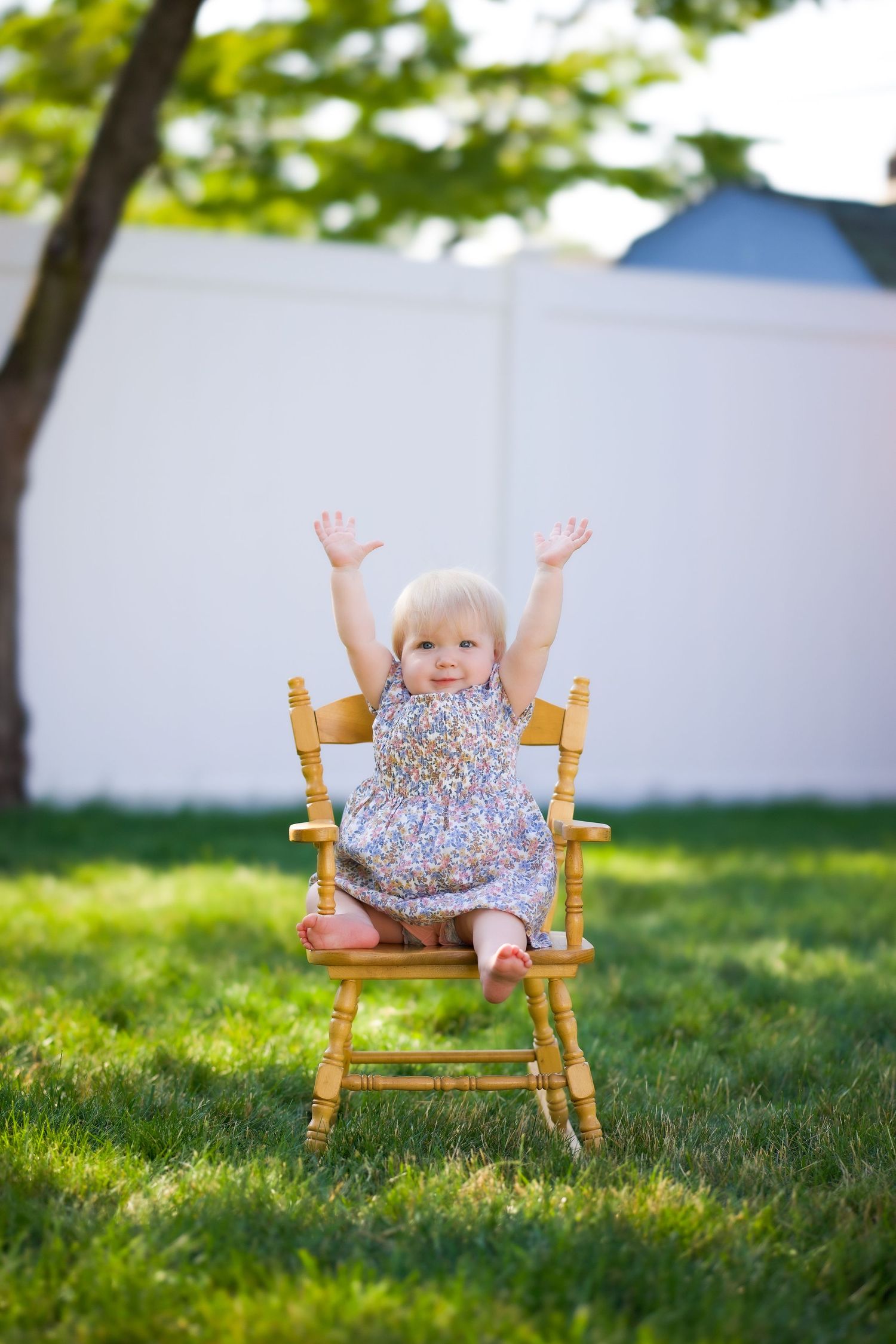 Small child sits in wooden rocking chair on green lawn at sunset with arms raised joyfully.