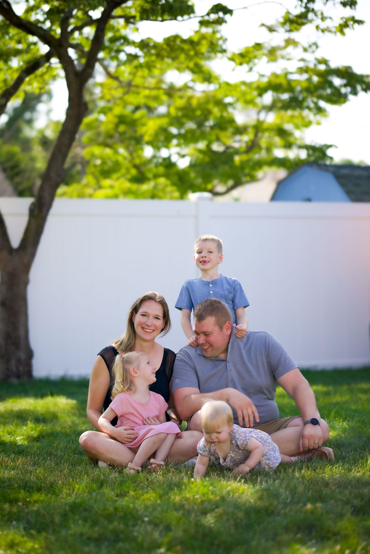 A family shares tender moments while laying on grass under leafy green trees.