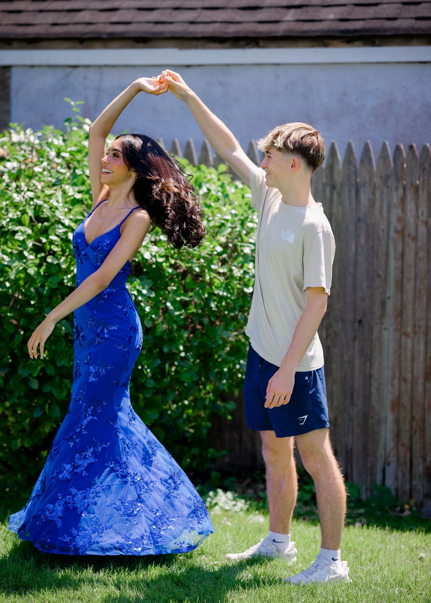 Two people dancing on a backyard lawn, one in a flowing blue mermaid dress and one in casual white and navy attire.