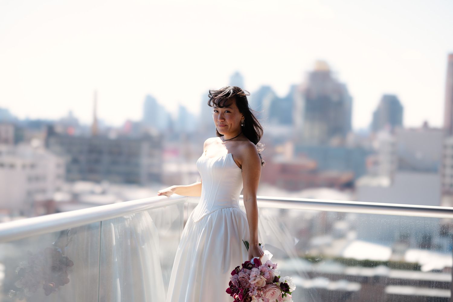 Bride in white dress holding purple bouquet on rooftop with city skyline backdrop.
