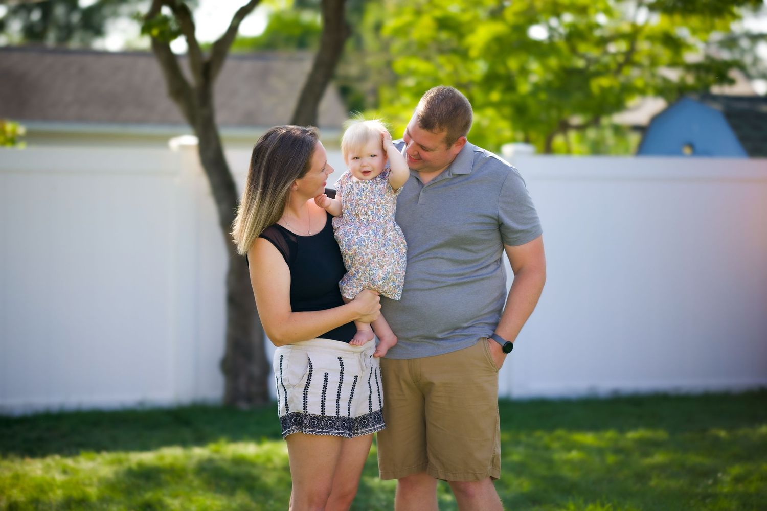 A family shares a joyful outdoor moment together on a sunny day in a backyard with a white fence and green trees.