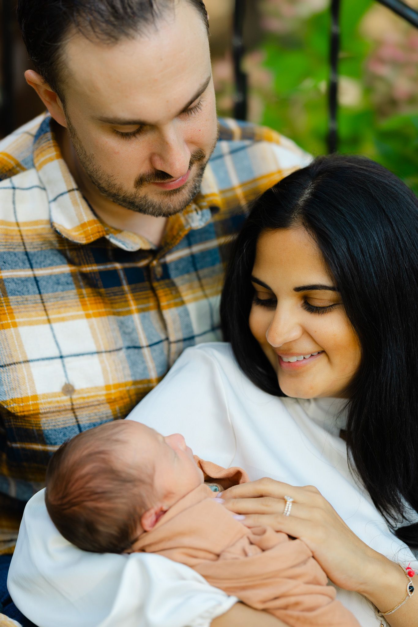 A couple gazes lovingly at their newborn baby while sitting outdoors in a natural setting.