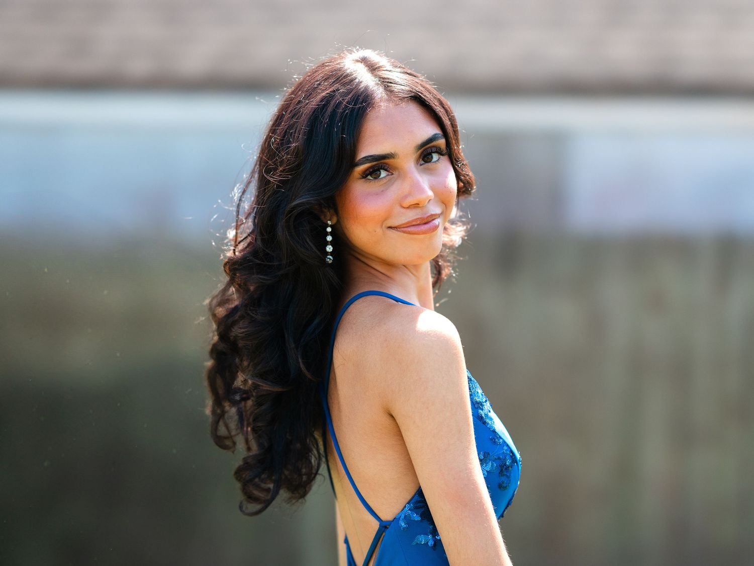 A woman with long dark curls wearing a blue evening dress smiles over her shoulder in soft natural lighting.
