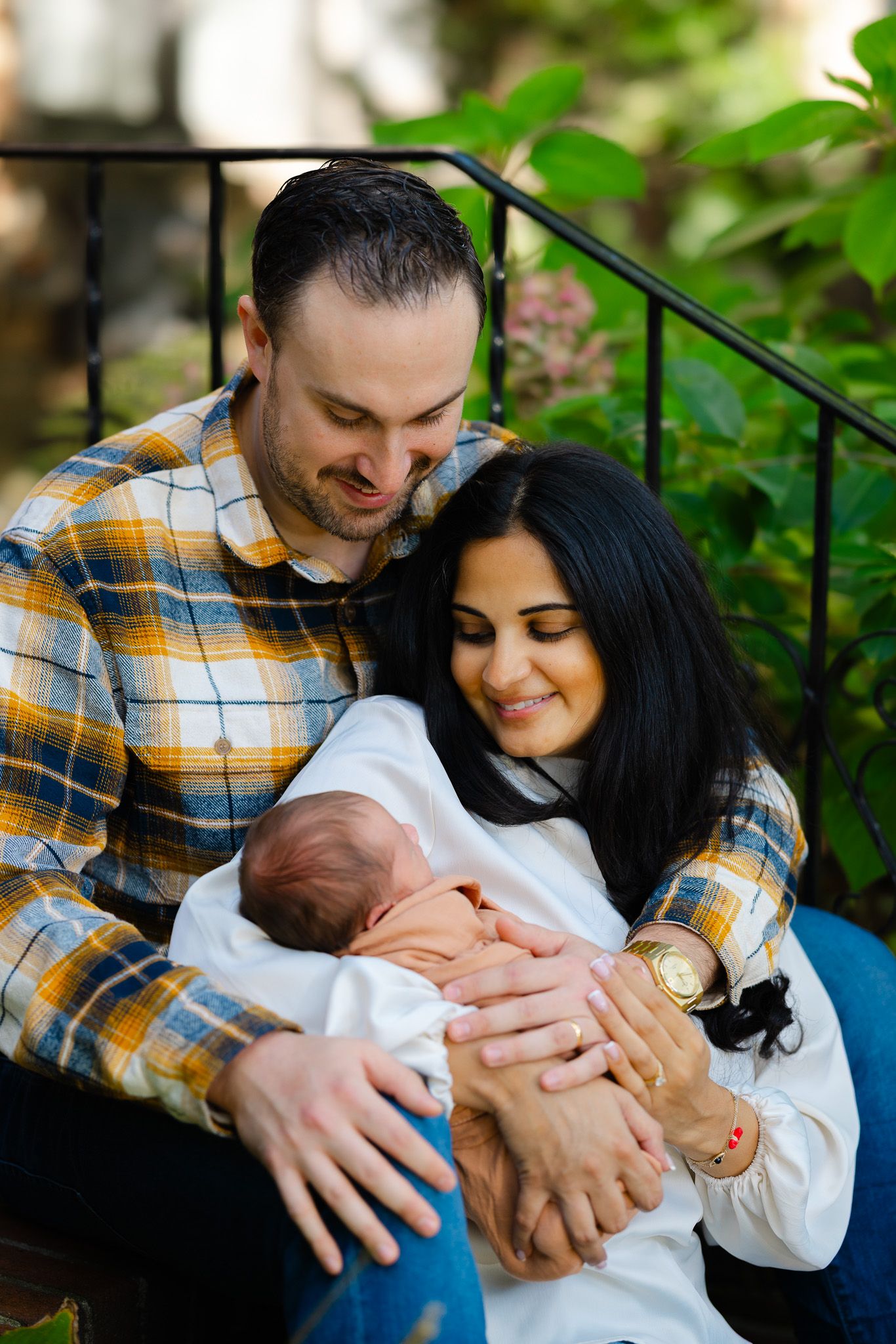 A couple shares tender moments with their newborn baby in a garden setting wearing casual attire.