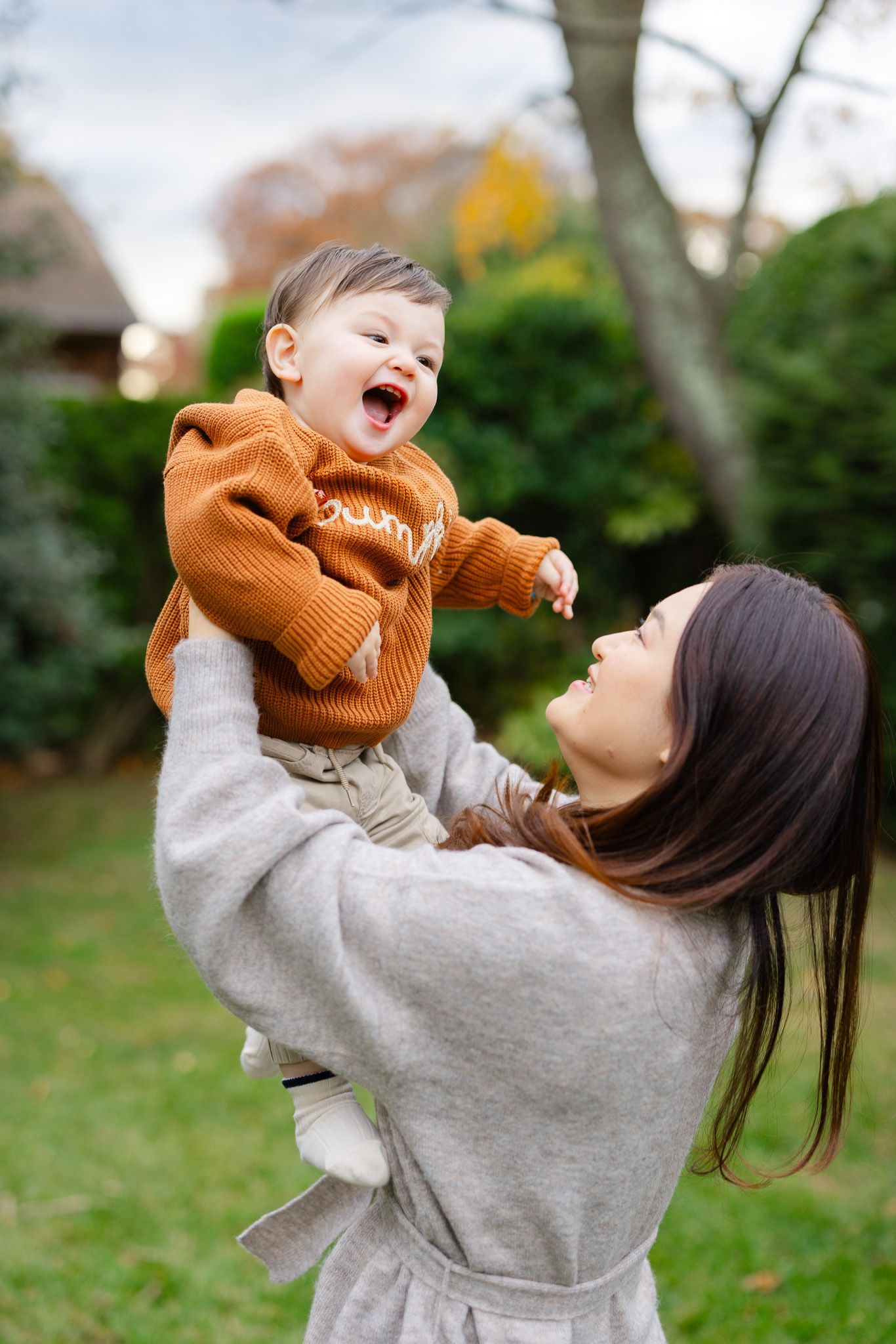 A joyful moment captured outdoors as a baby in an orange sweater is lifted up in the air during playtime.