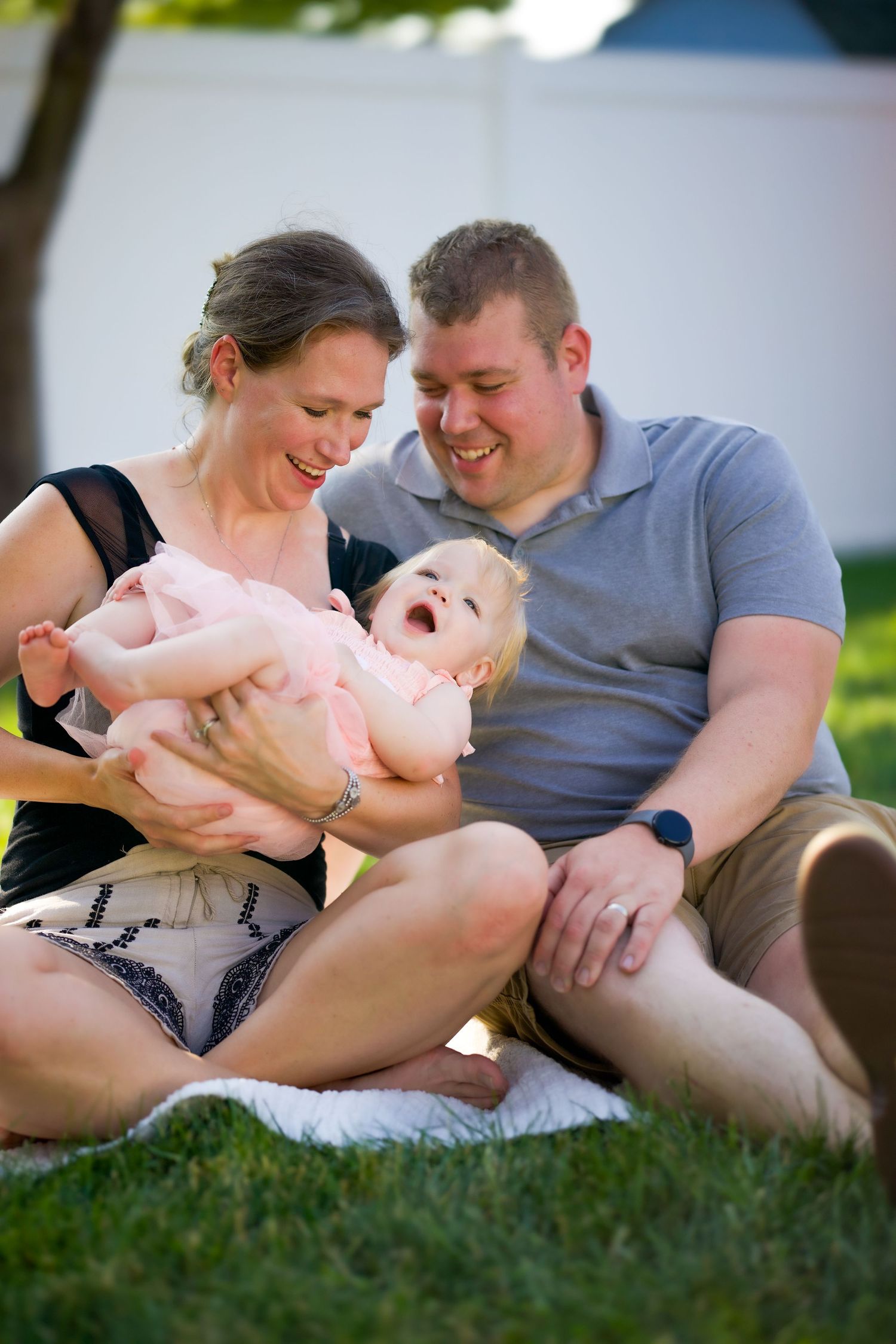 A happy family shares a joyful moment together while sitting on grass during a sunny outdoor photoshoot.