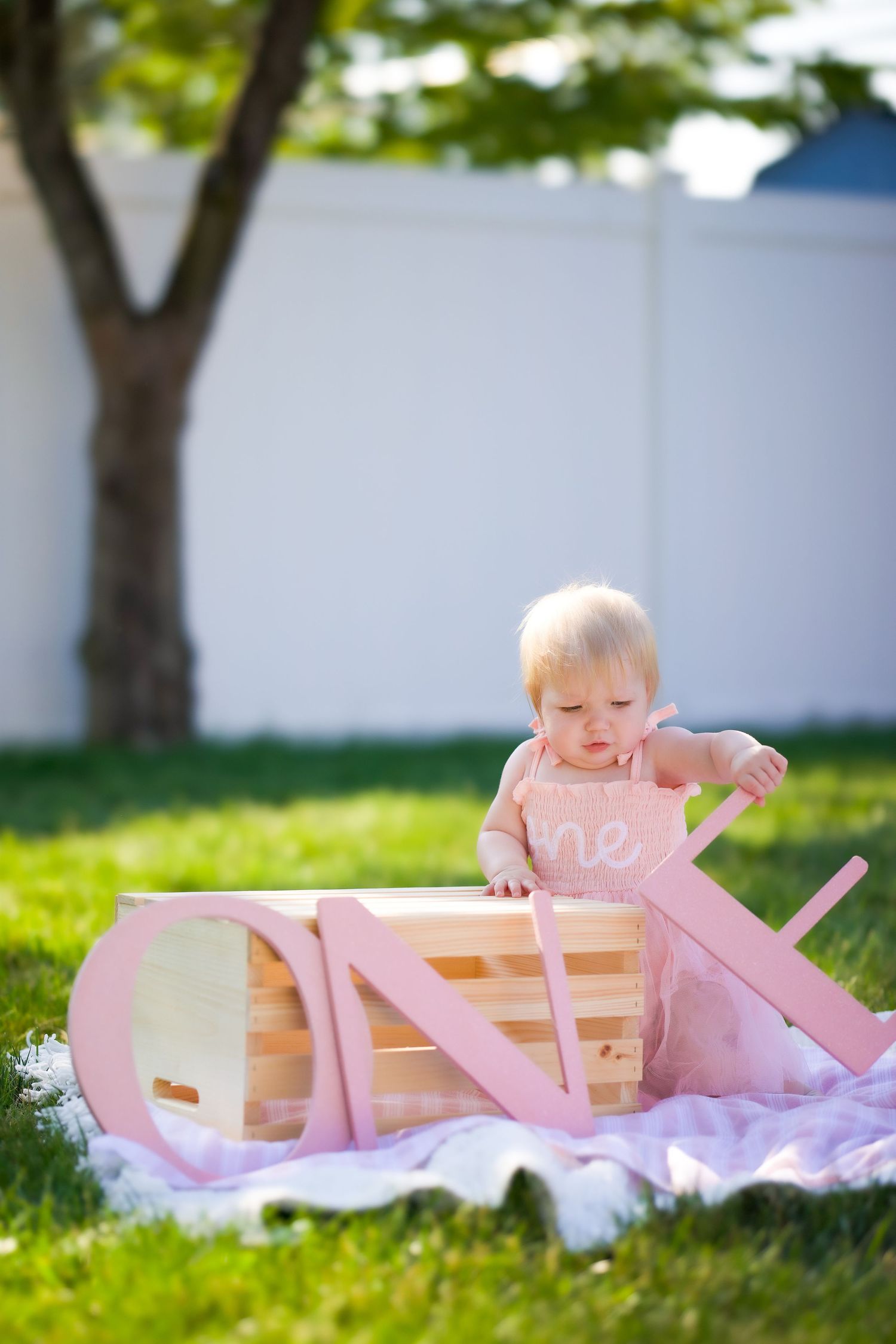 A small child in pink plays with a wooden rocking boat on grass in a sunny backyard setting.