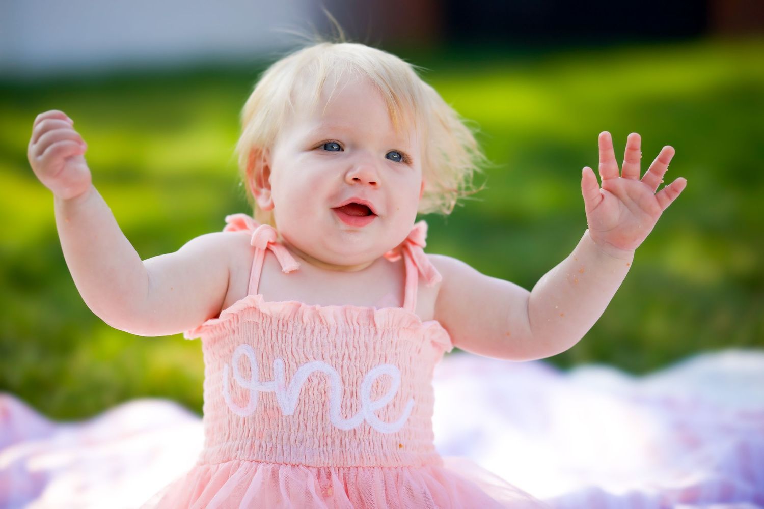 A toddler in a pink dress raises their hands with joy while playing outdoors on a sunny day.