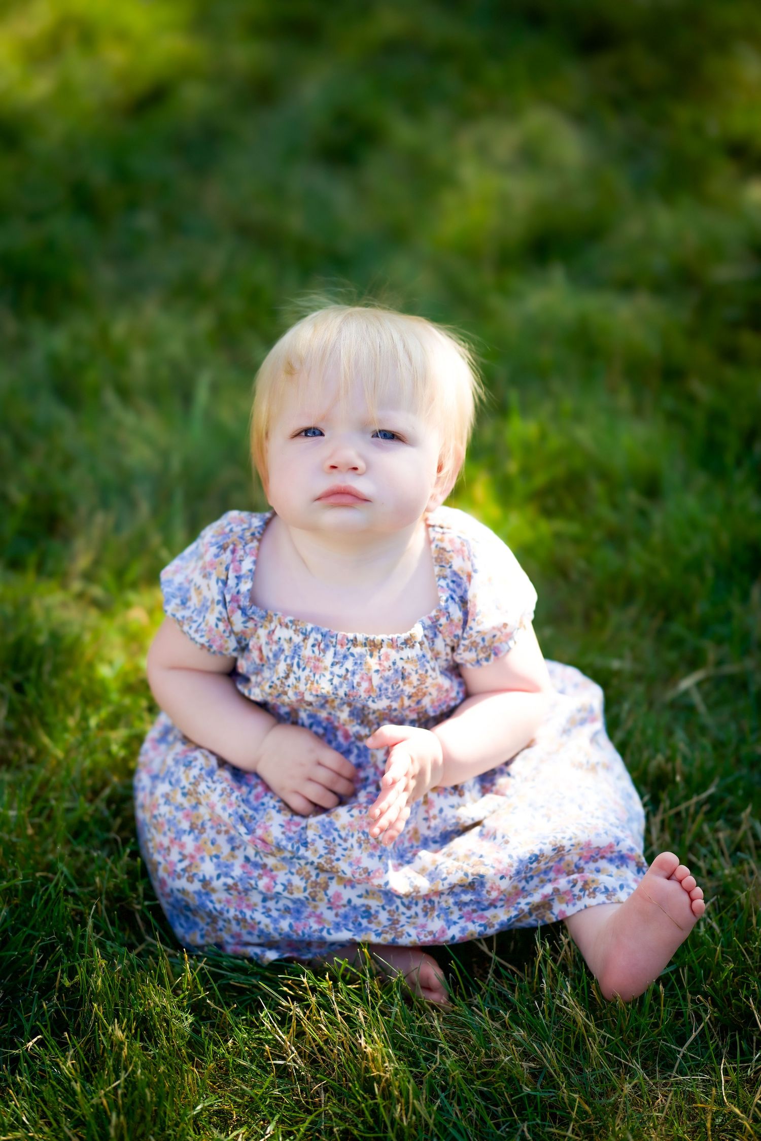 A small child sits on green grass wearing a floral patterned dress with short puff sleeves.