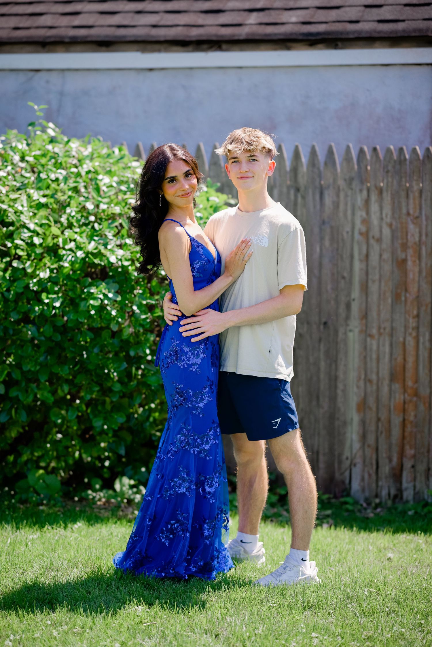 A couple poses outdoors in a backyard, with one person wearing a sparkly blue formal gown and the other in casual attire.