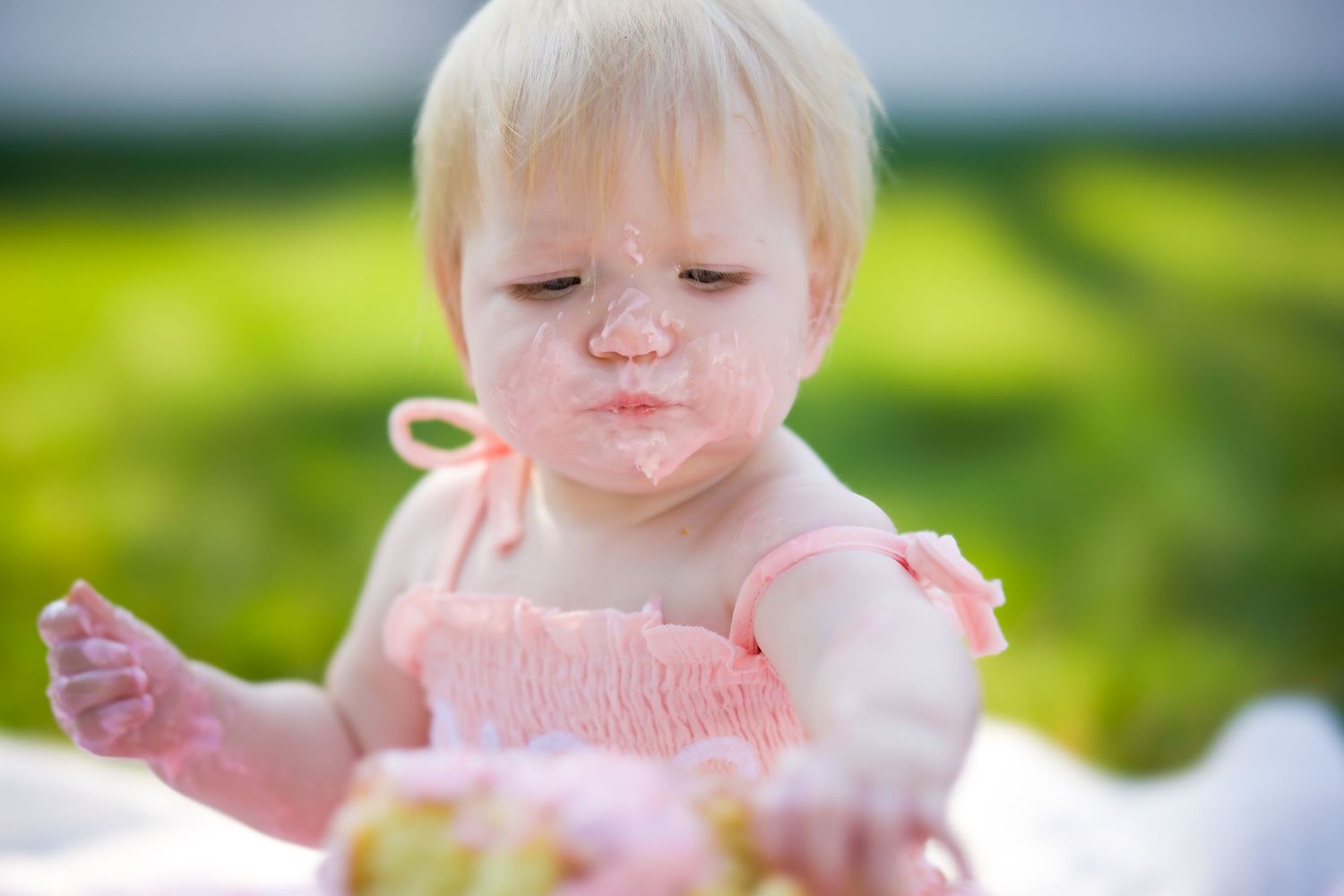Baby dressed in pink smiling outdoors in bright sunlight with green grass backdrop.