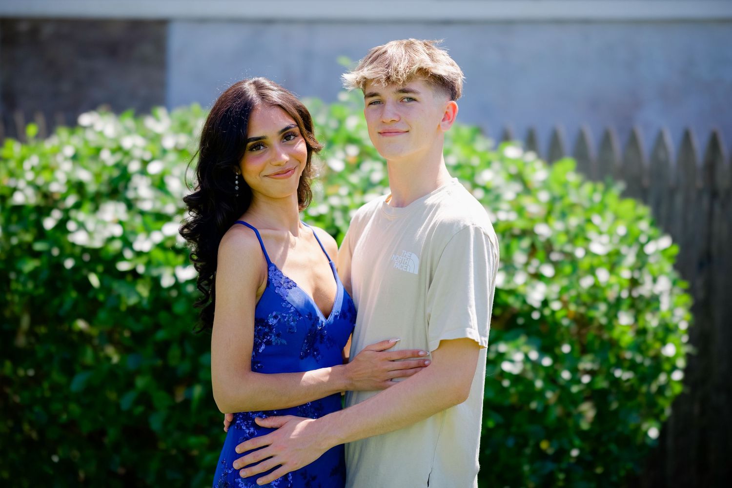 A couple in royal blue and white embraces outdoors in a garden setting with lush green foliage as backdrop.
