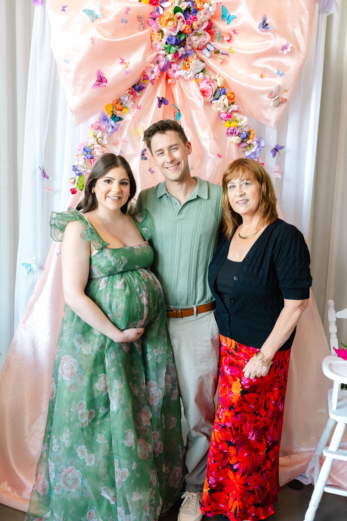 Three people pose in front of a pink backdrop decorated with colorful butterflies and floral elements at a celebration.