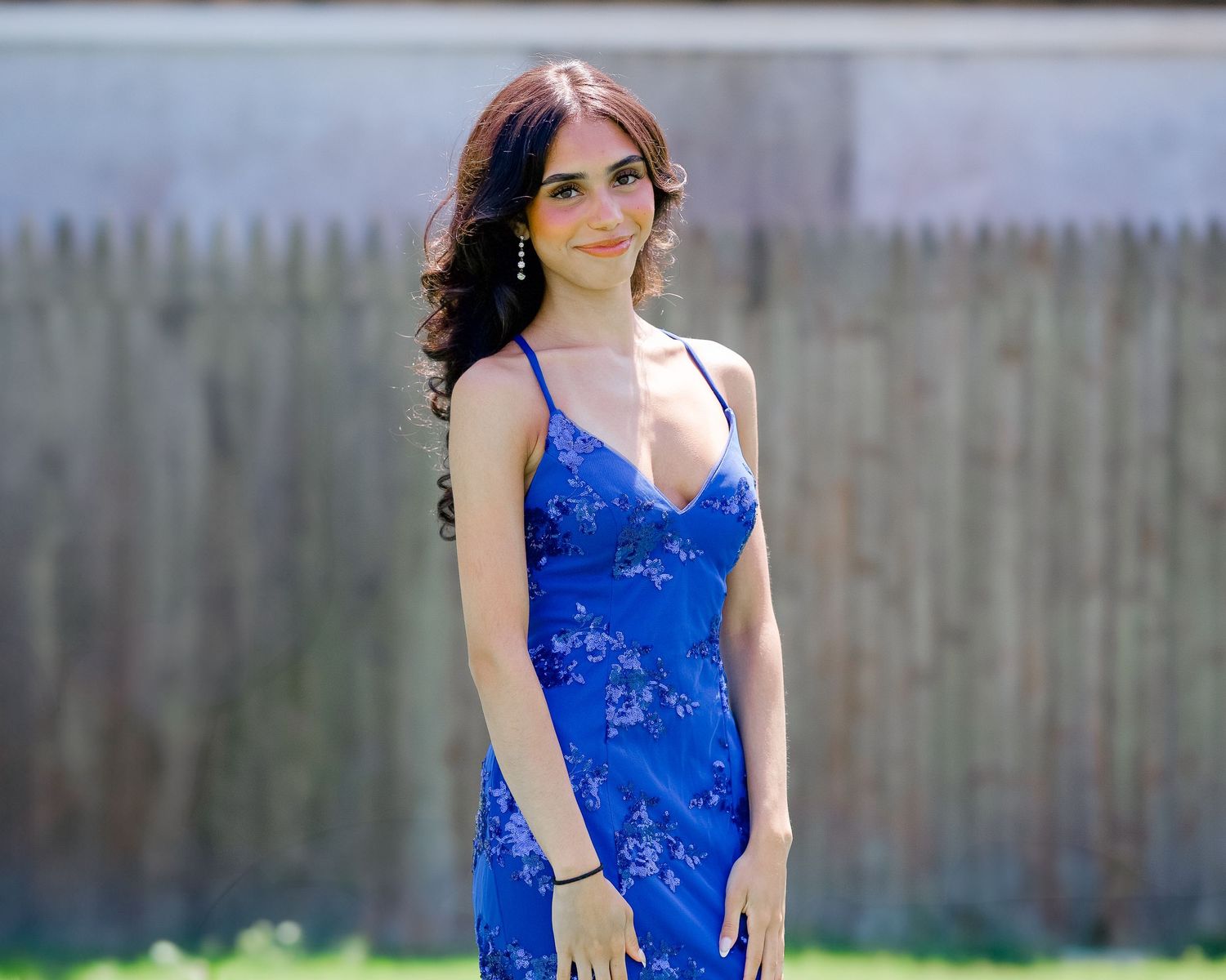Woman in a royal blue formal dress poses outdoors in front of wooden fence.