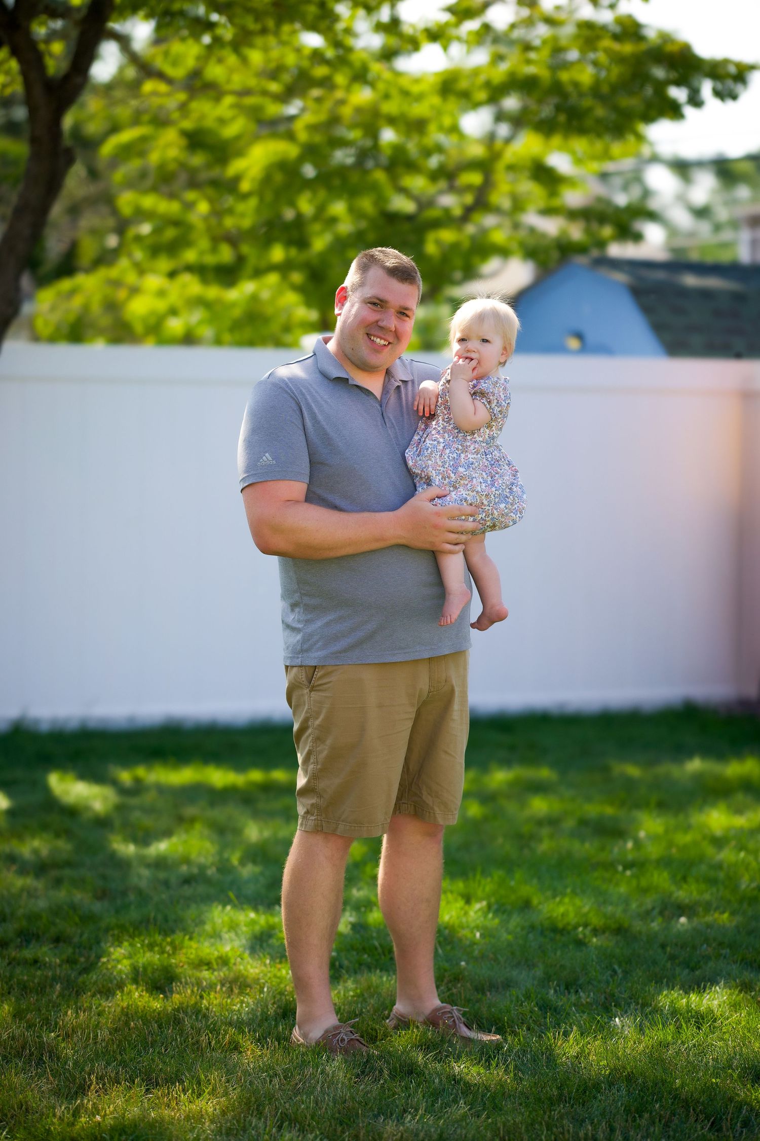 On sunny grass, an adult in gray shirt and khaki shorts holds a small toddler in a floral dress as they share a playful moment.