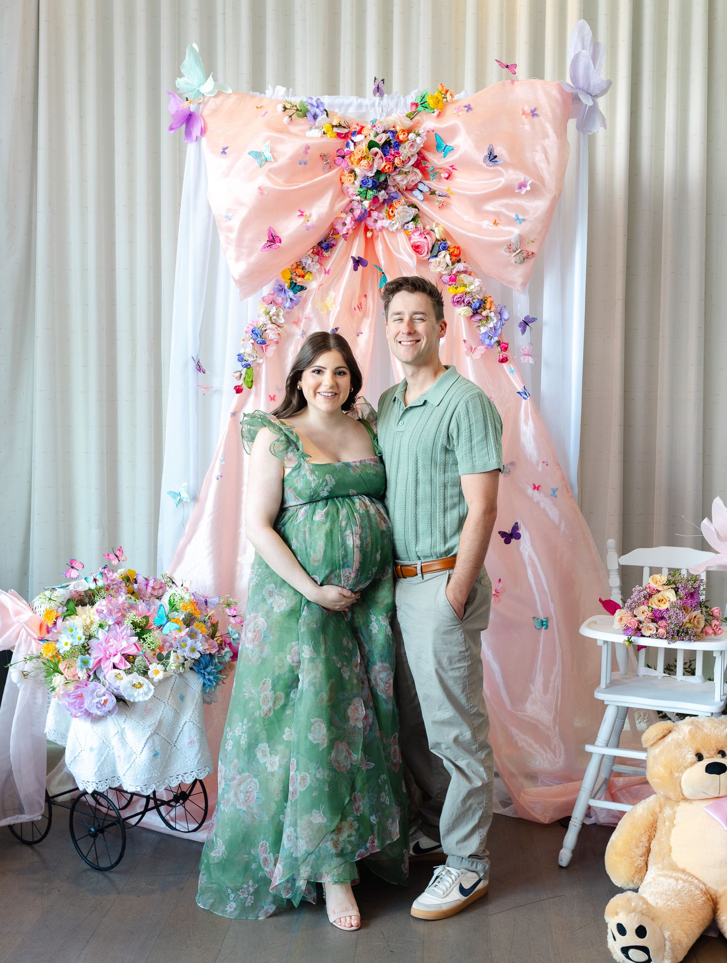 Baby shower celebration setup with pink and floral decorations against white backdrop featuring green dress attire.