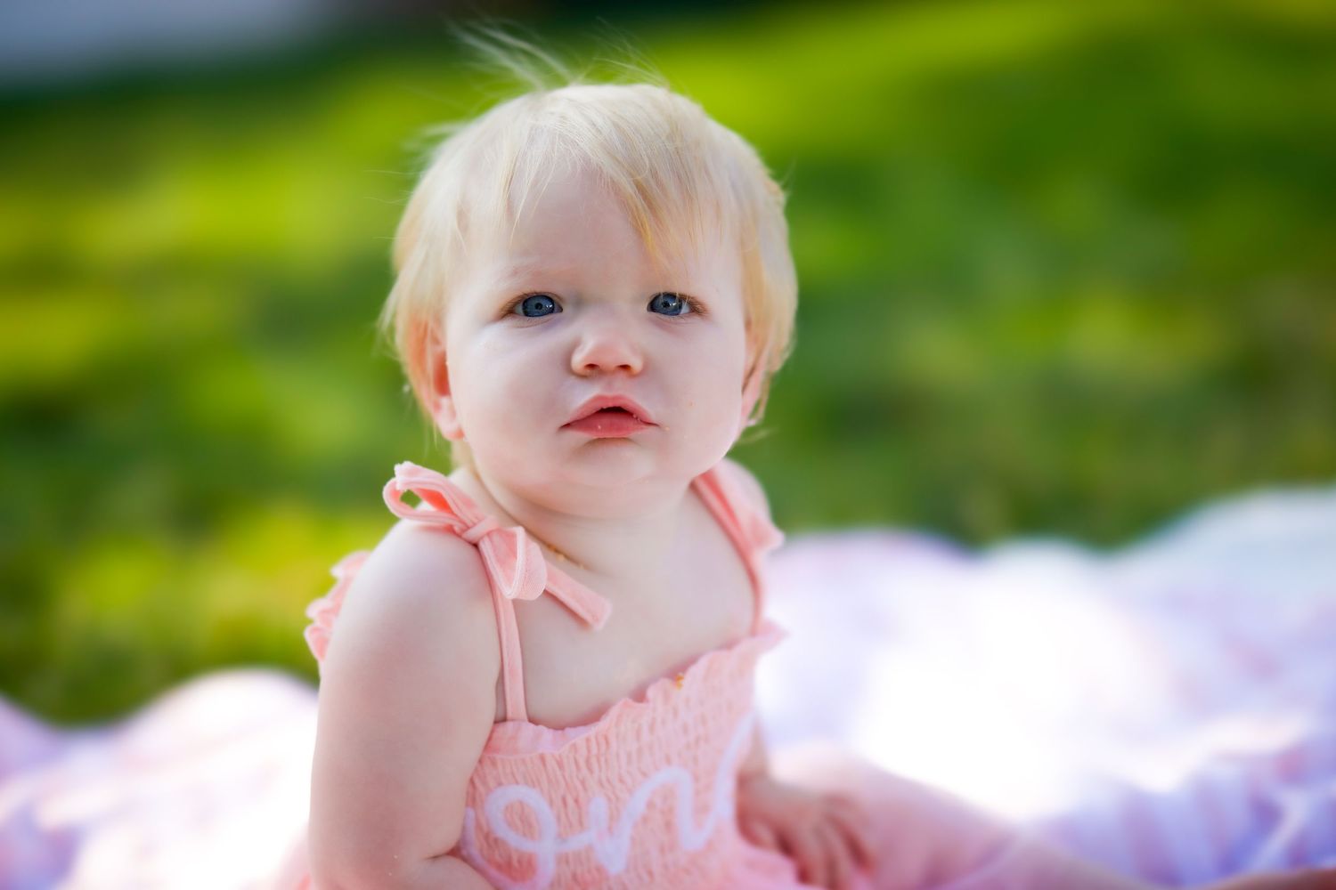 A baby in soft pink clothing rests outdoors against a blurred green nature background.