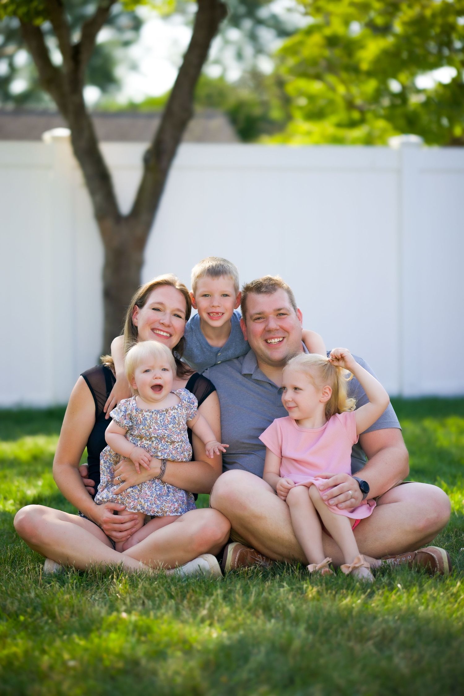 A family sits together and laughs while relaxing on green grass in their backyard on a sunny summer day.