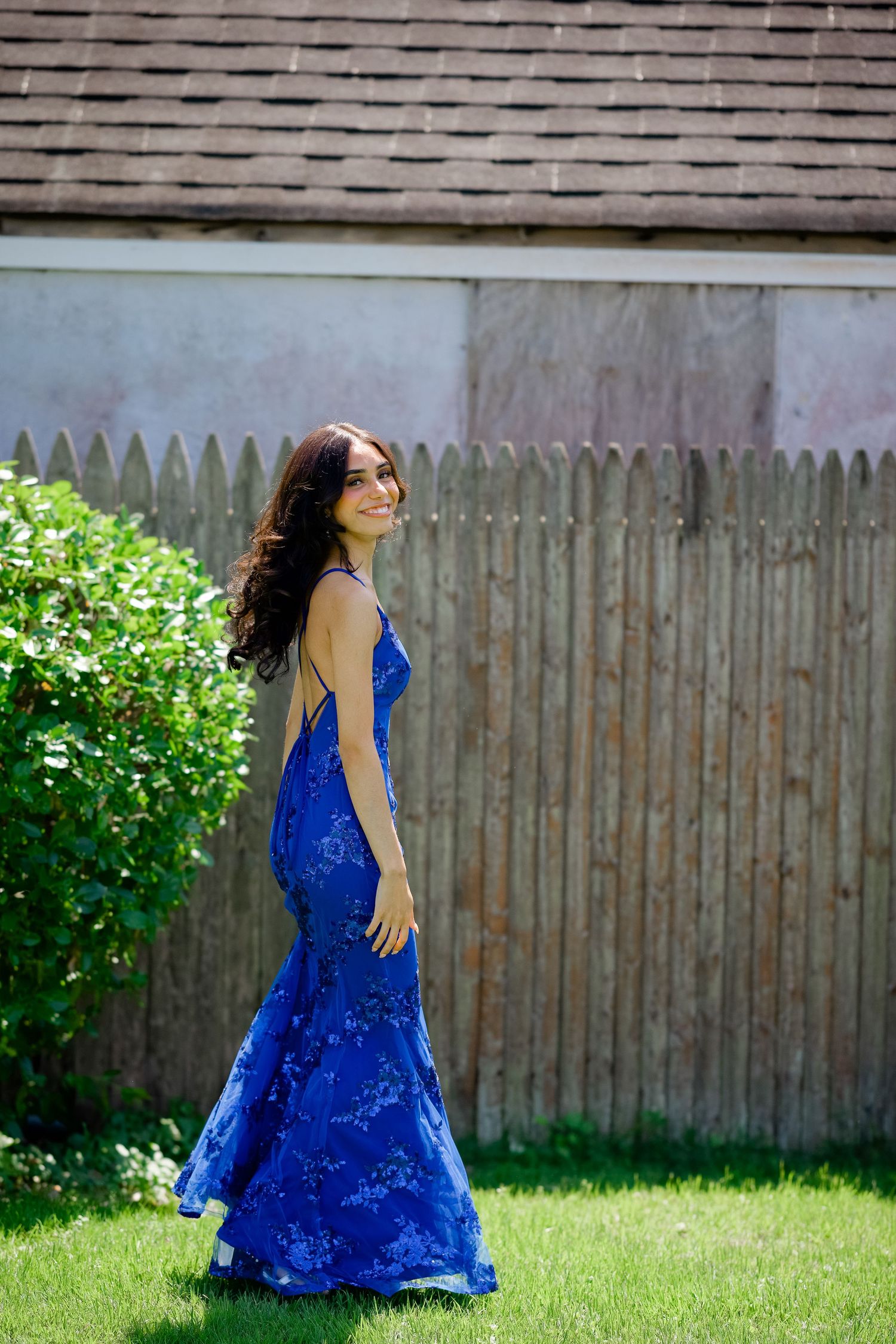 A person in a flowing royal blue dress twirls and dances gracefully in a sunny backyard with a wooden fence.