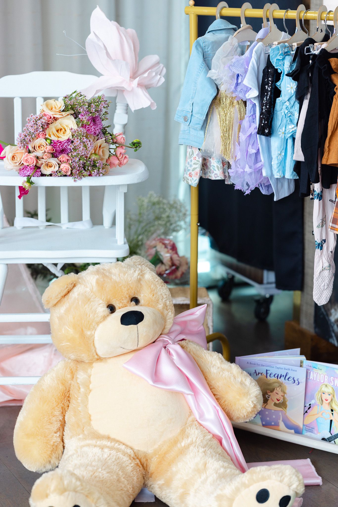 Soft plush teddy bear with pink bow displayed among baby clothes and nursery decor in a pastel-themed setting.
