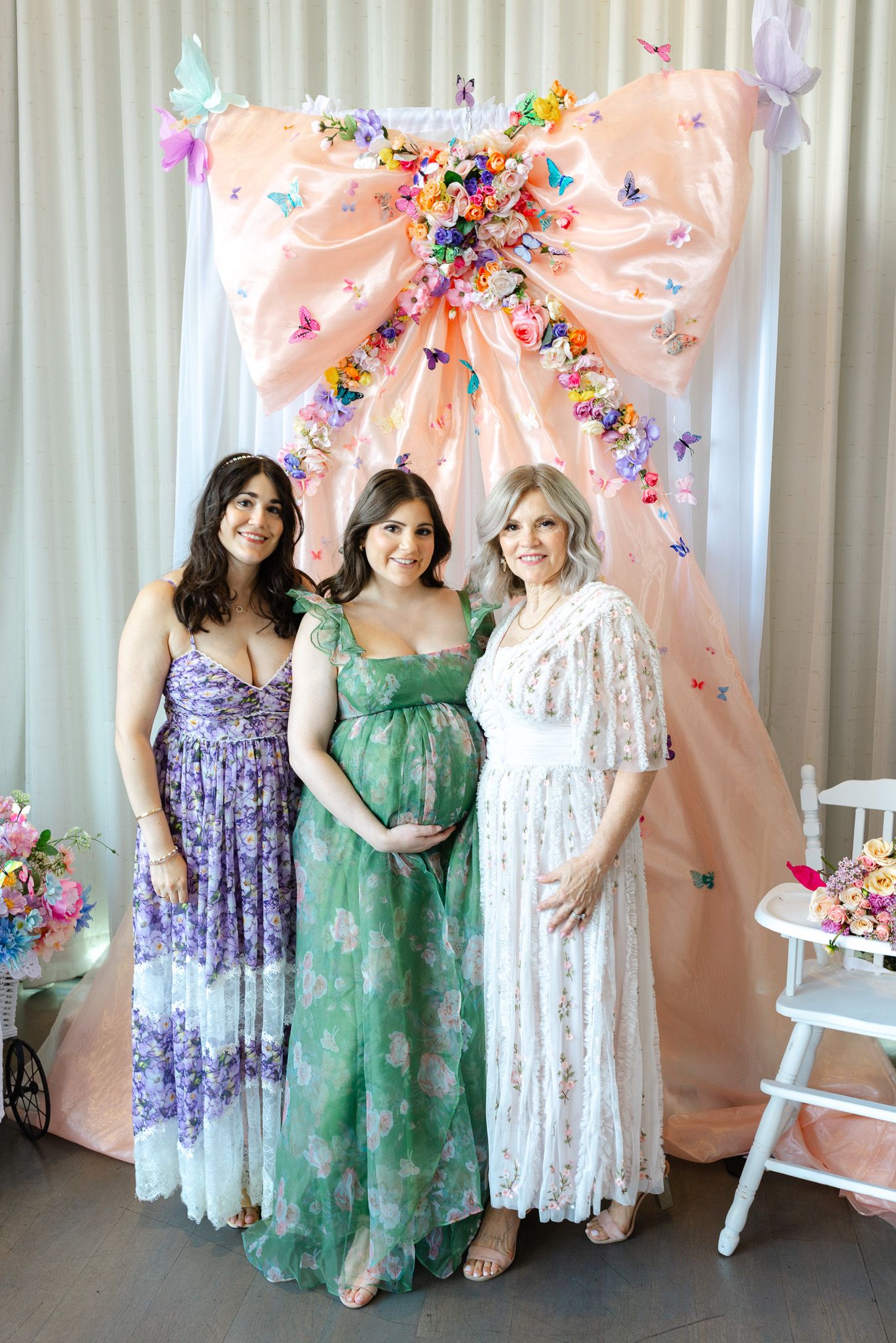 Group photo at a baby shower celebration with floral decorations and pink bows against a white backdrop.