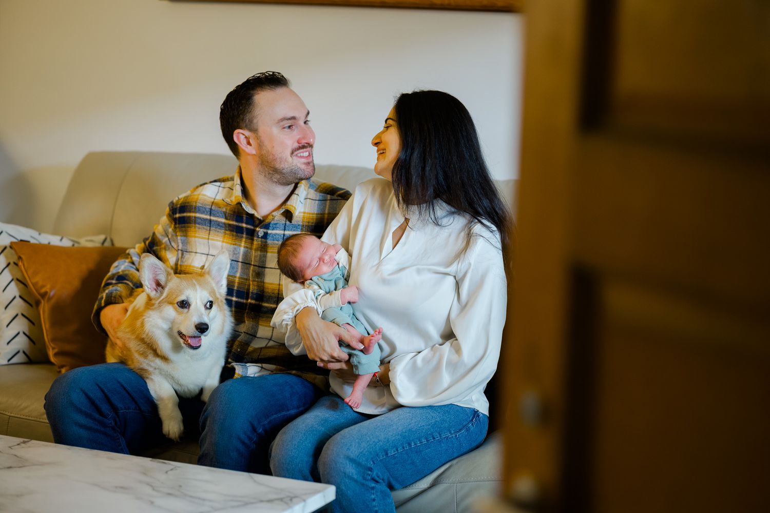 A couple sits on a brown leather couch with their Corgi dog during an indoor photoshoot session.