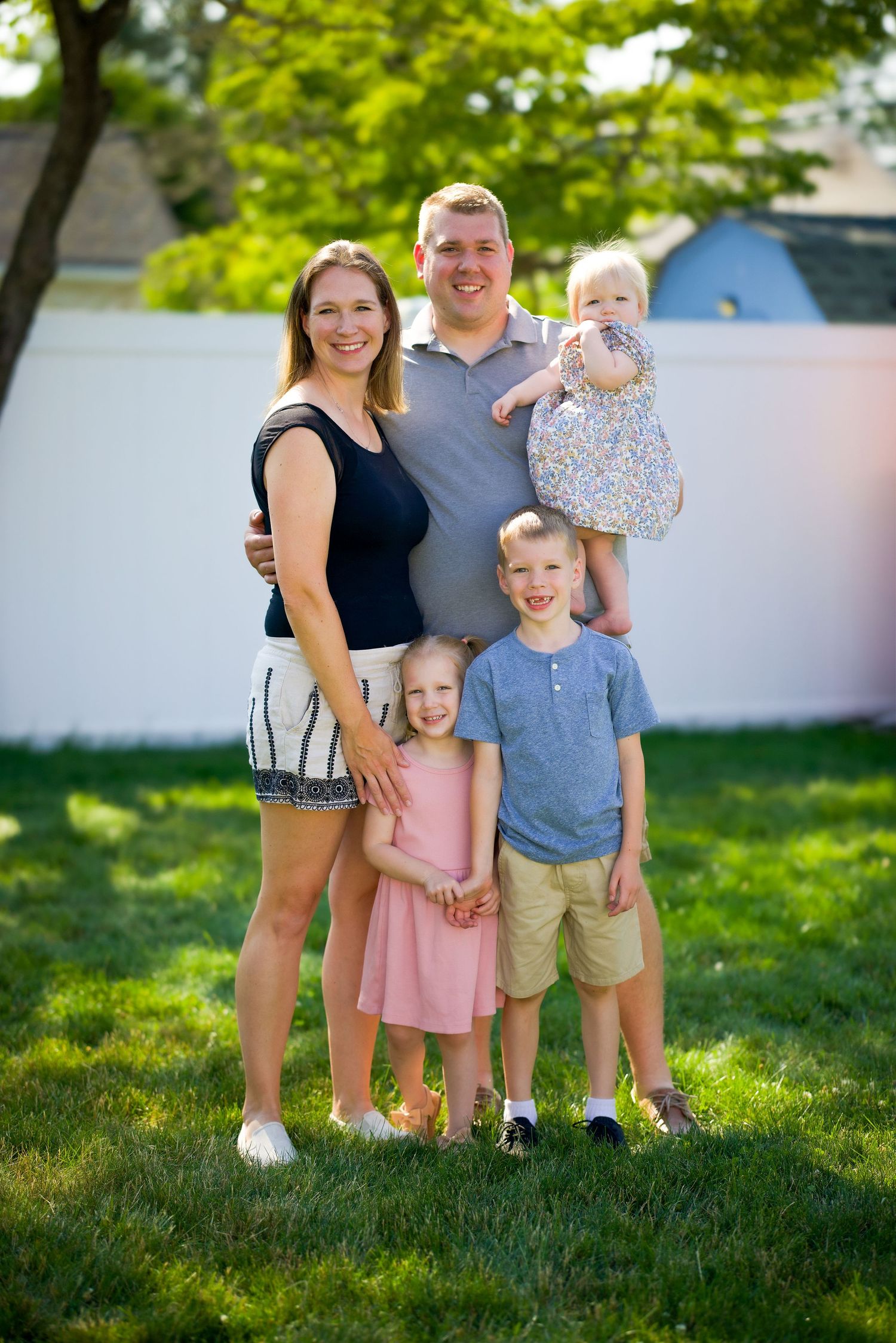 A family stands together on a sunny summer day with green trees and a white wall in the background.