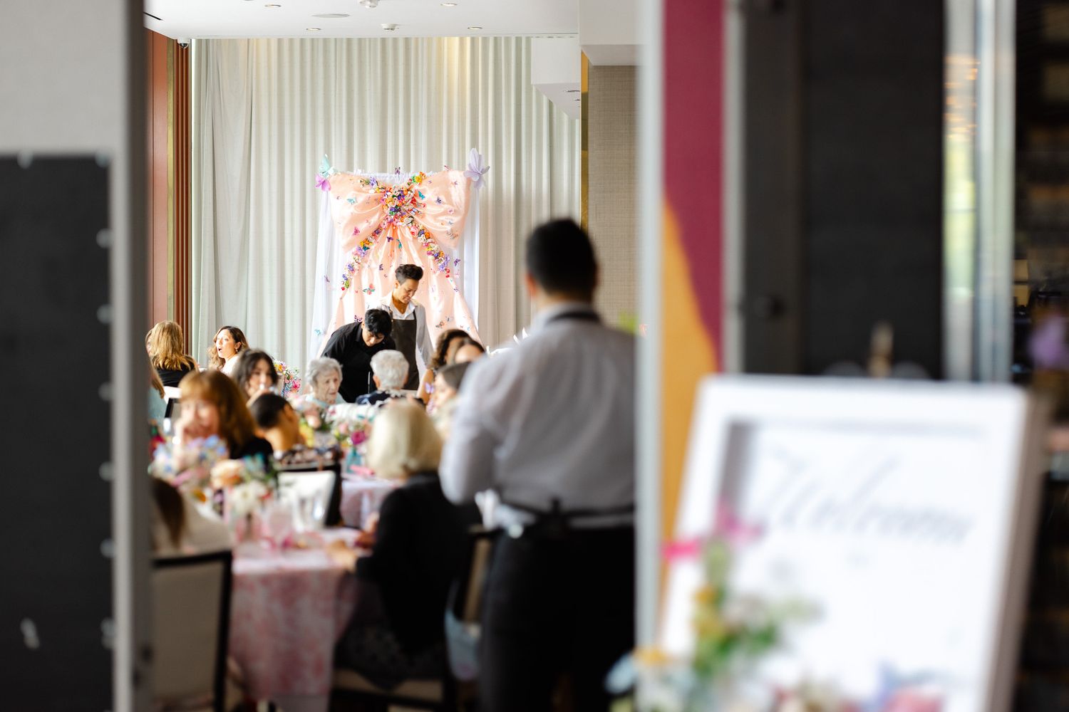 A wedding reception venue with white curtains, tables, and guests mingling in an elegant indoor setting.
