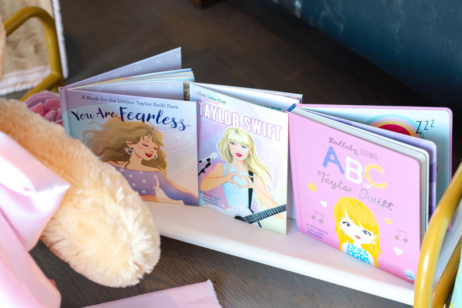 Pink and pastel colored children's books are displayed on a shelf with soft plush toys.