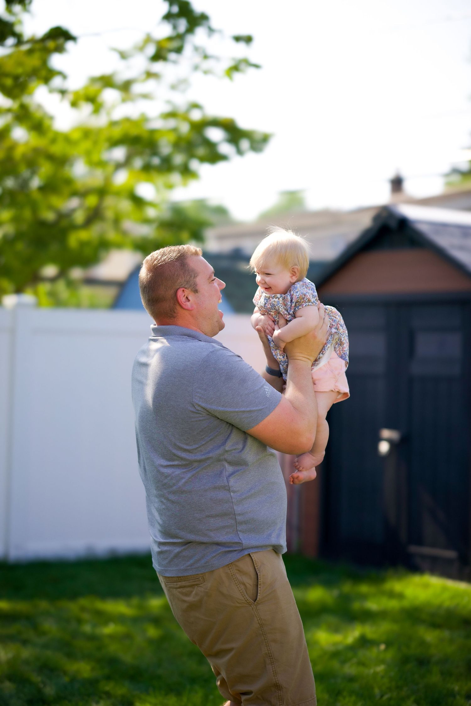 A father holds and lifts his baby in a backyard setting with a shed and green foliage in the background.