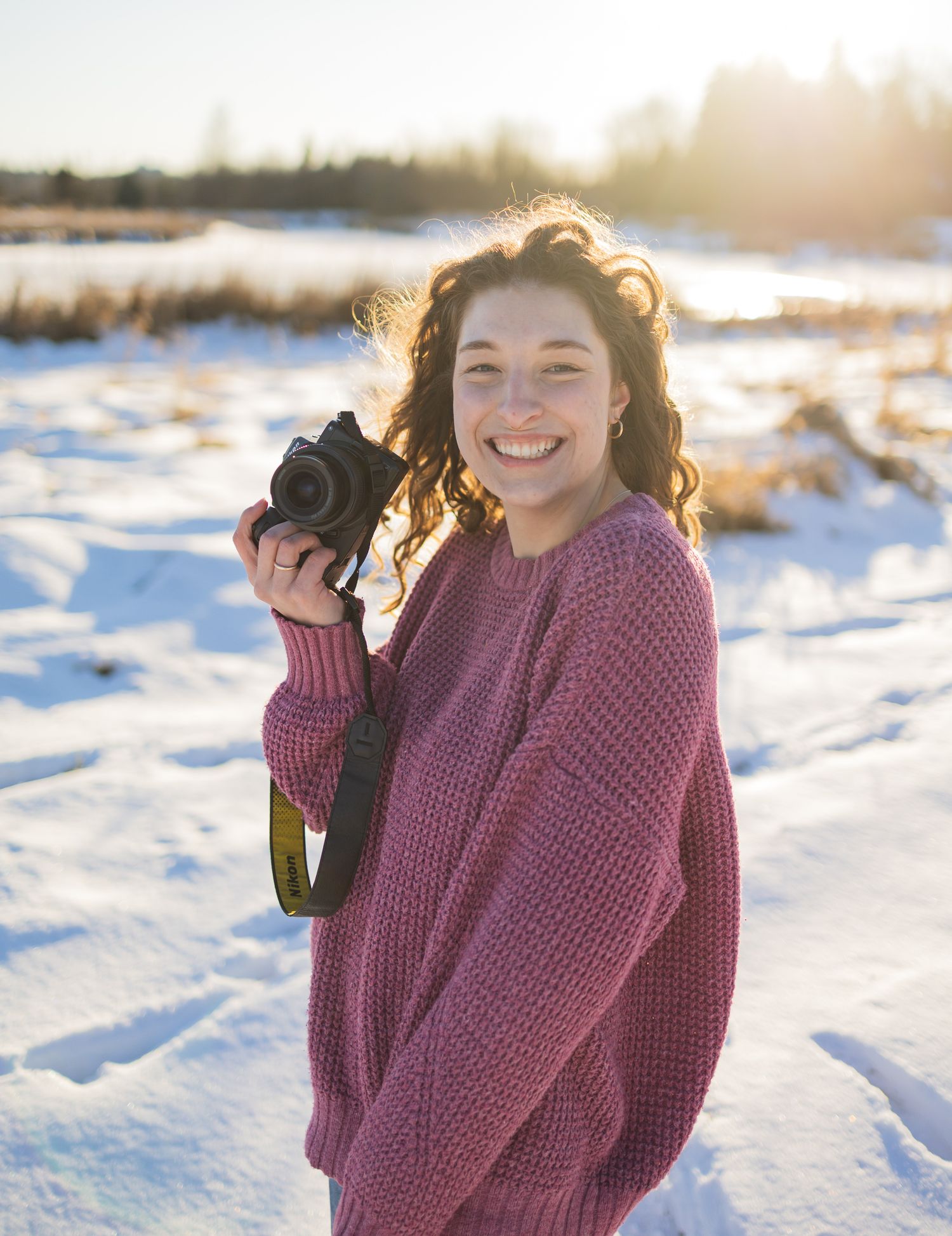 a woman in a pink sweater, smiling with her camera in the snow.