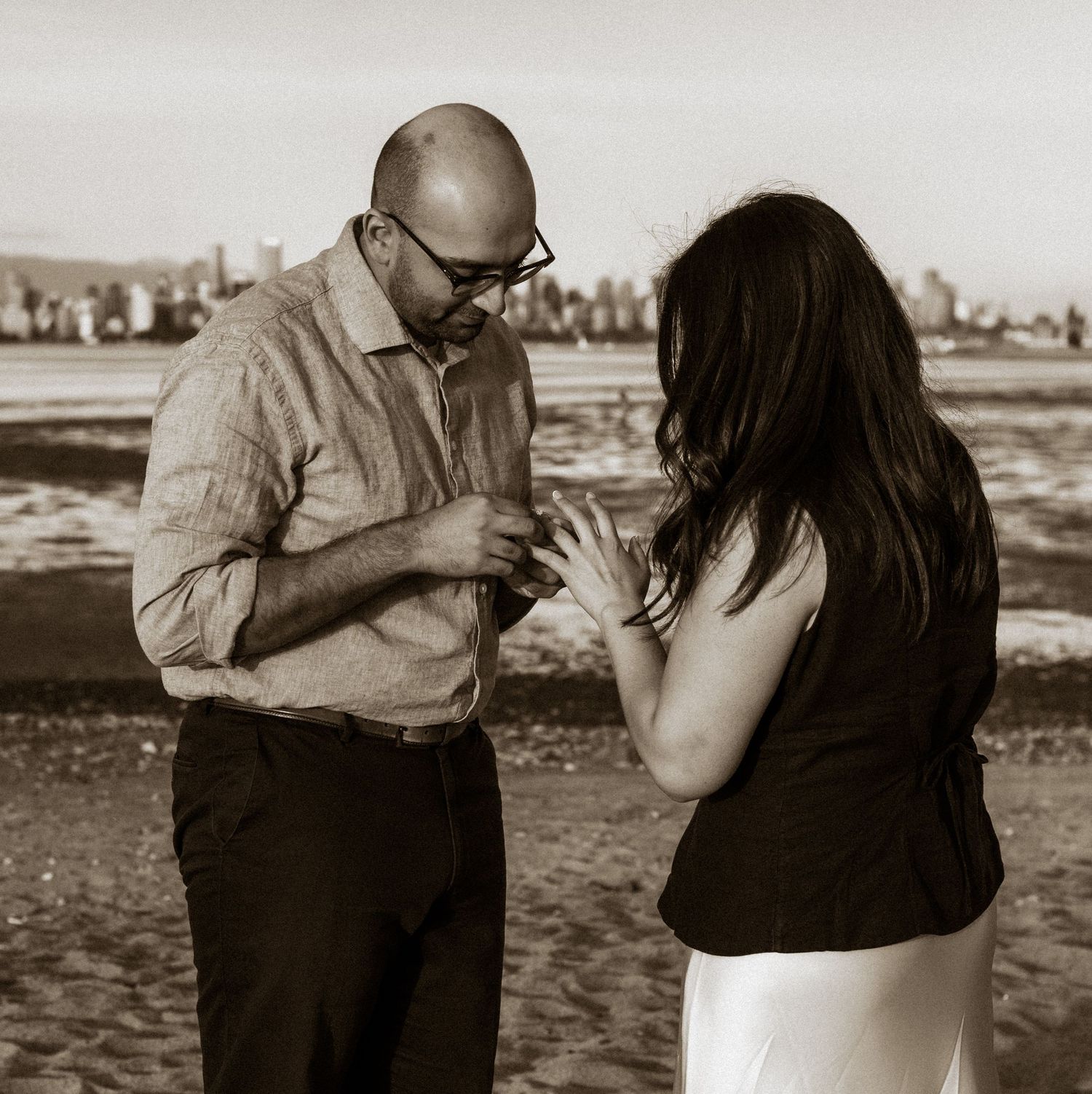 man putting ring on womans finger on the beach with city view in the background
