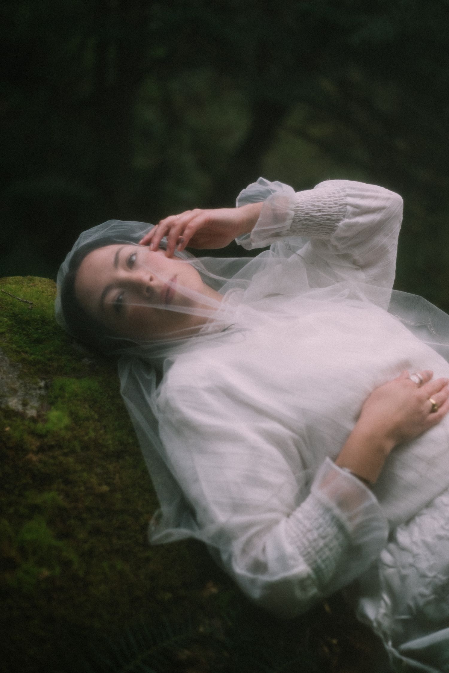 A dreamy, close-up portrait of a woman with dark hair, partially veiled, lying on mossy ground in a natural forest setting.