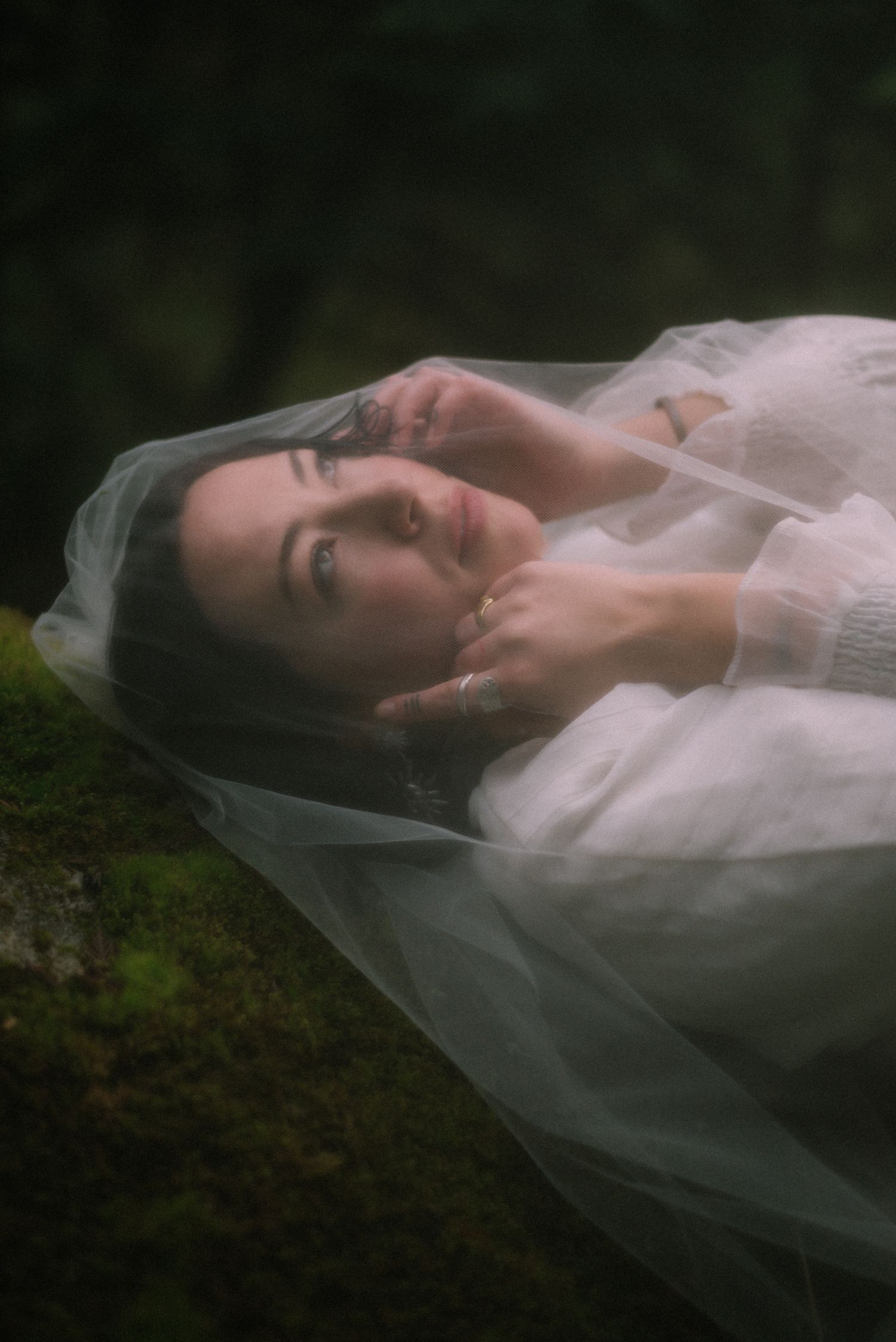 A dreamy, close-up portrait of a woman with dark hair, partially veiled, lying on mossy ground in a natural forest setting.