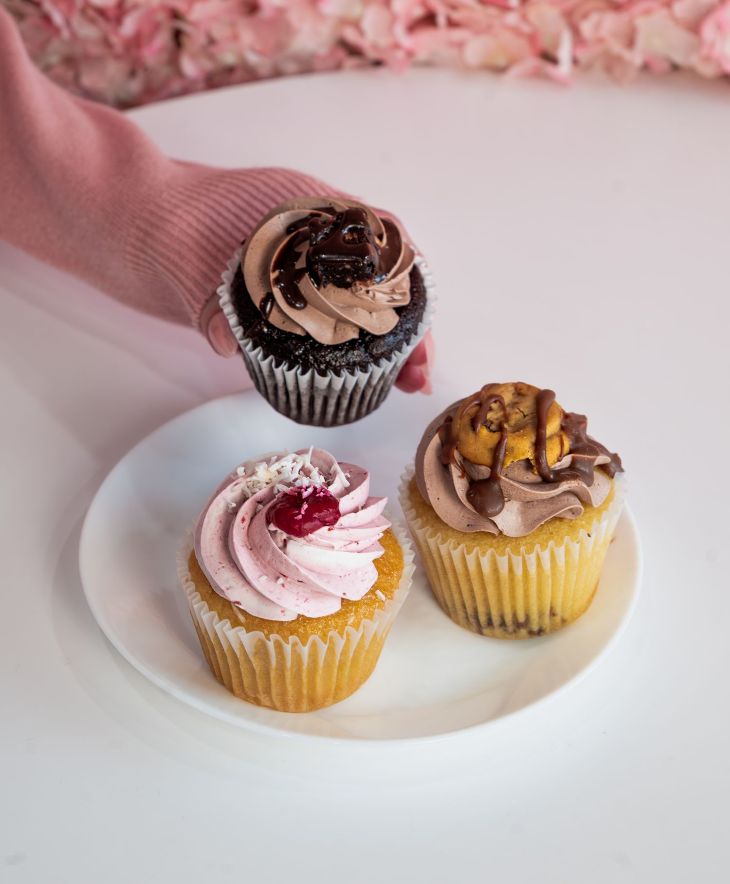 Three beautifully decorated gourmet cupcakes on a white plate, with a hand reaching for a chocolate cupcake.