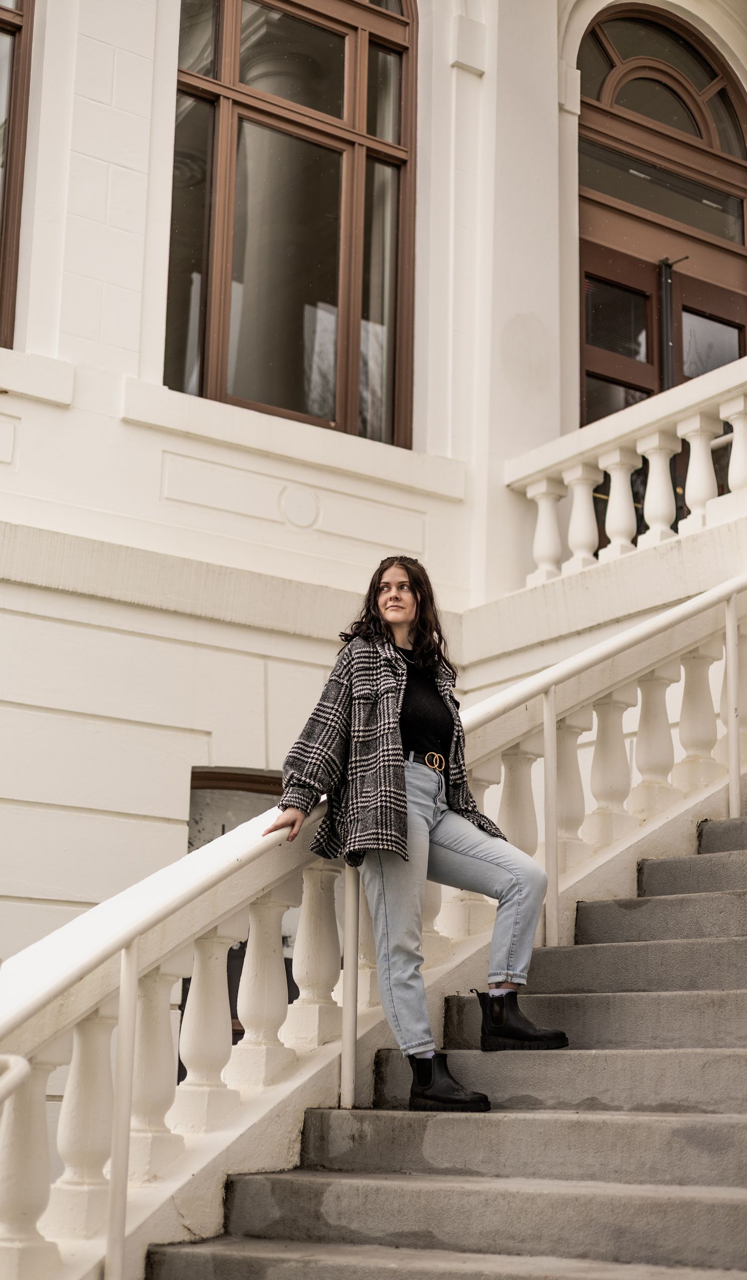 A young woman with dark, curly hair wearing a plaid jacket and jeans poses on the concrete steps with white railings of a white building