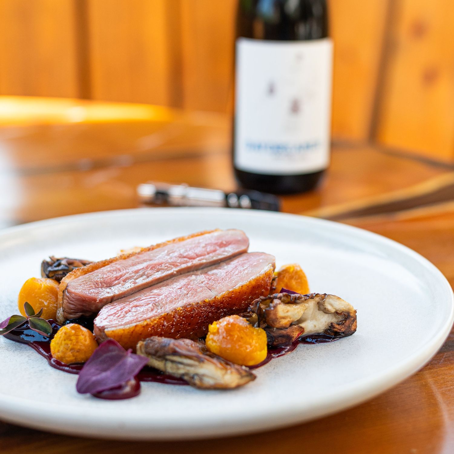 a colourful plate of duck breast with vegtables and wine in the ackground