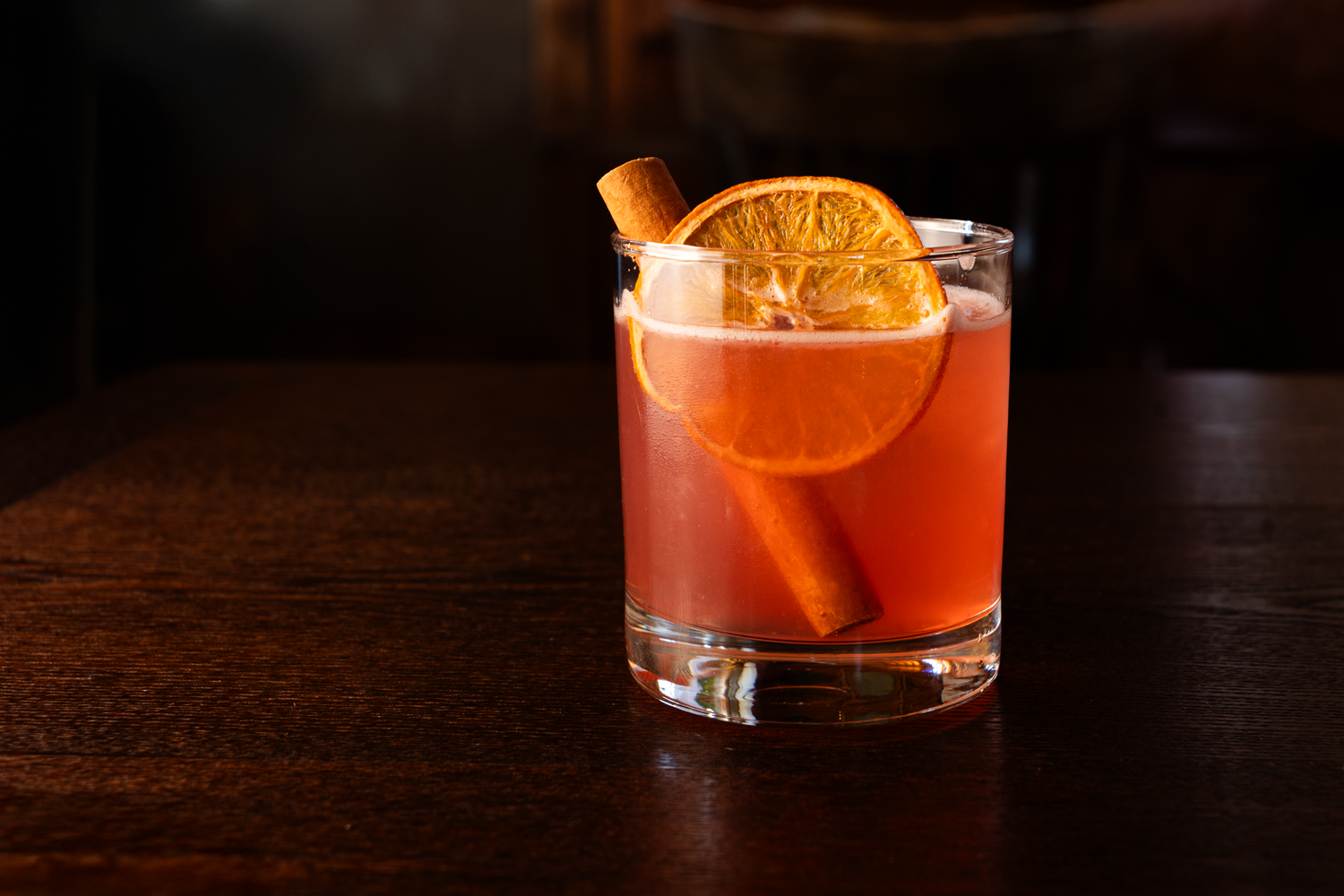 A vibrant orange cocktail in a clear glass, garnished with a dried orange slice and a cinnamon stick, sitting on a dark wooden surface.