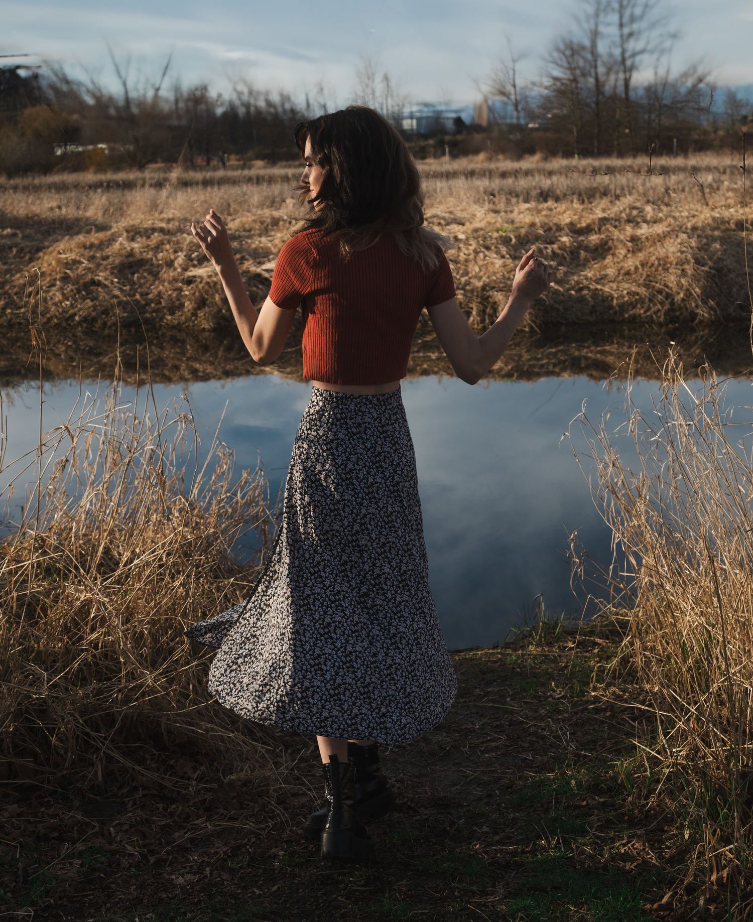 Woman by River in Fraser Valley.
A woman with dark, wavy hair, wearing a cropped rust-colored top and a floral print skirt.