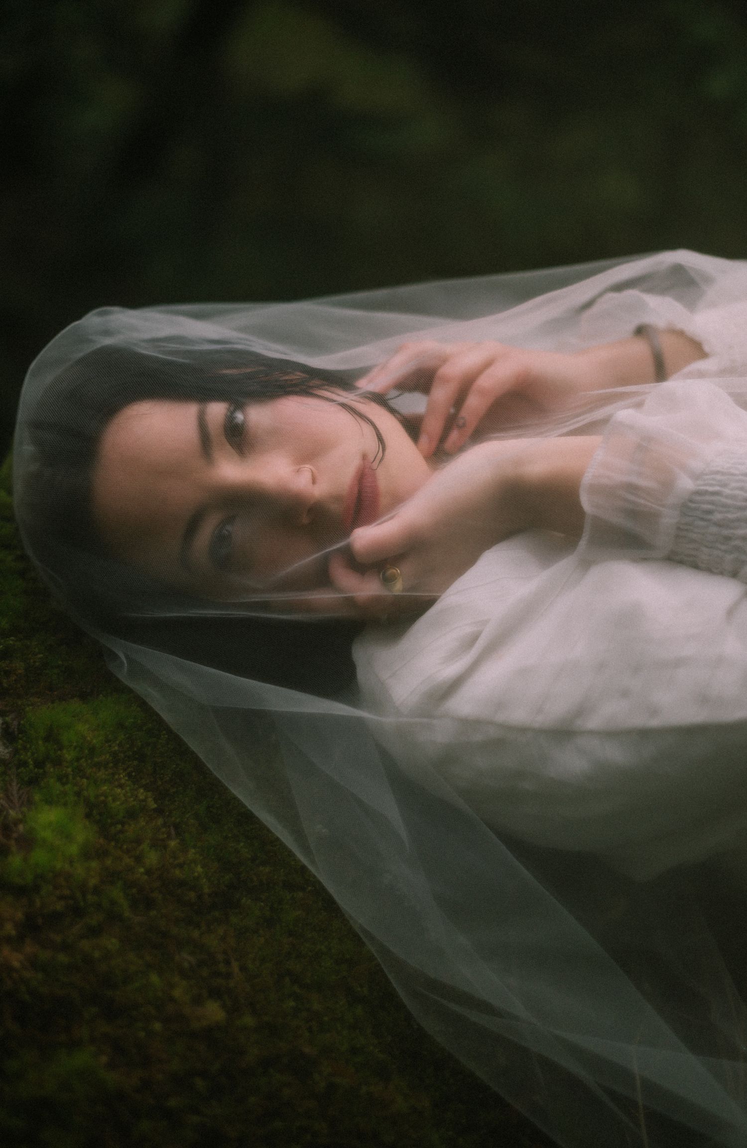 A dreamy, close-up portrait of a woman with dark hair, partially veiled, lying on mossy ground in a natural forest setting.