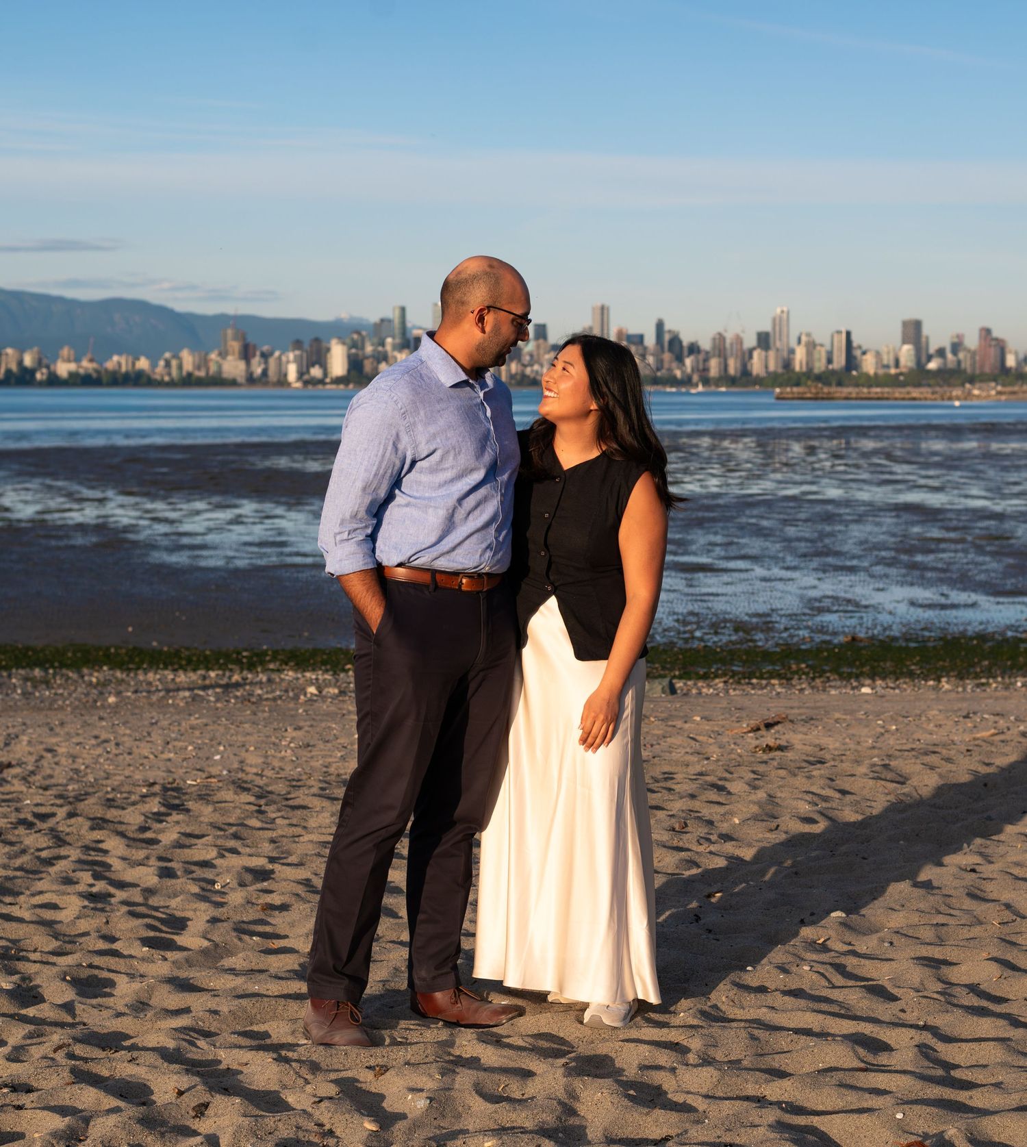 man and woman smiling at eachother on the beach with city view in the background