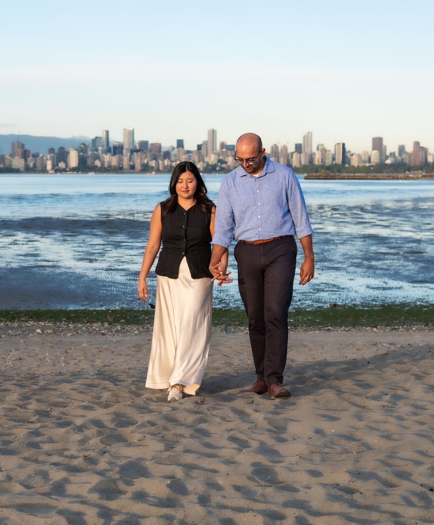 man and woman hand in hand walking on the beach with city view in the background
