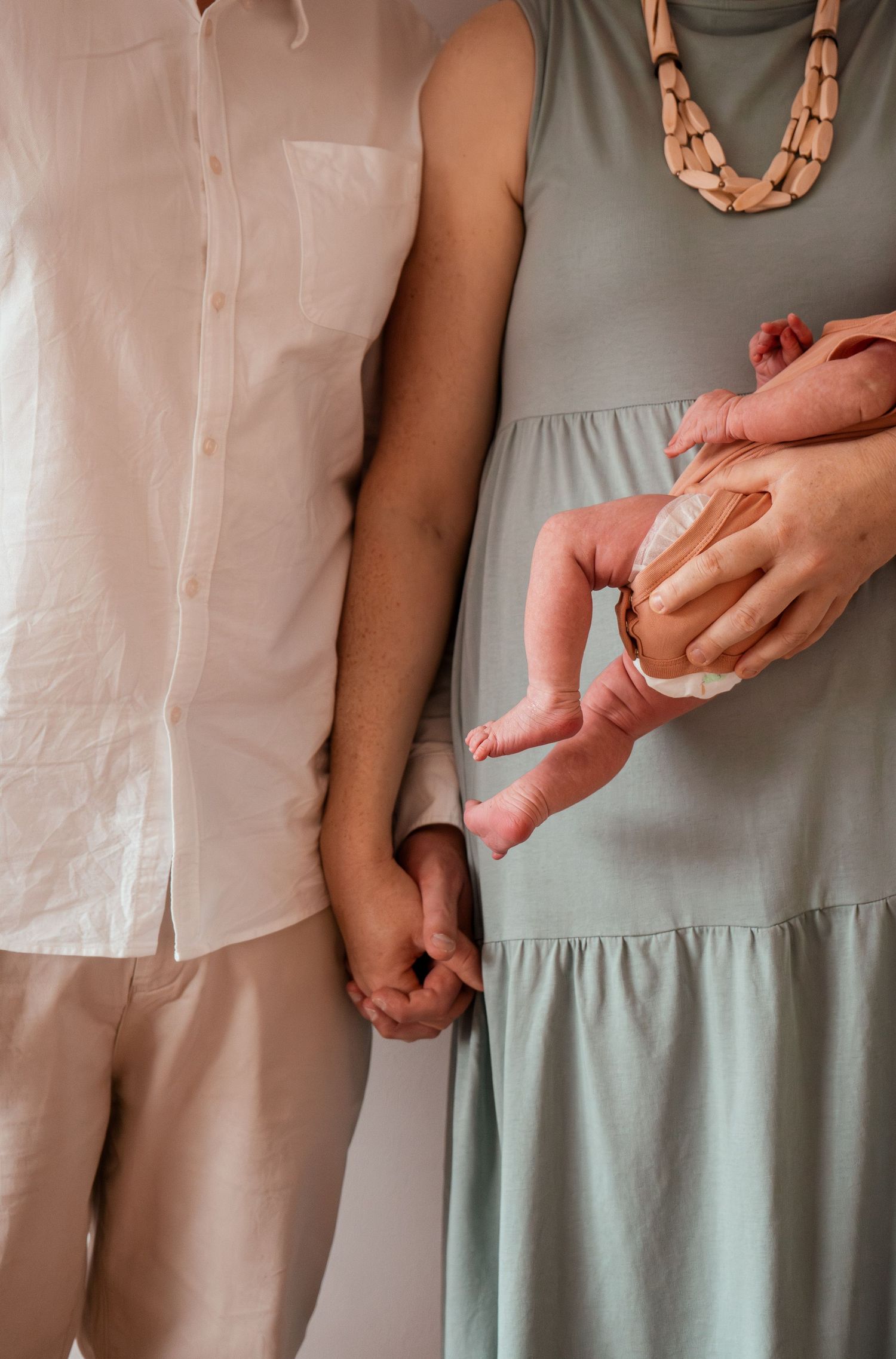 Close-up of hands holding a newborn baby, with a sage green dress and white shirt visible in the frame.